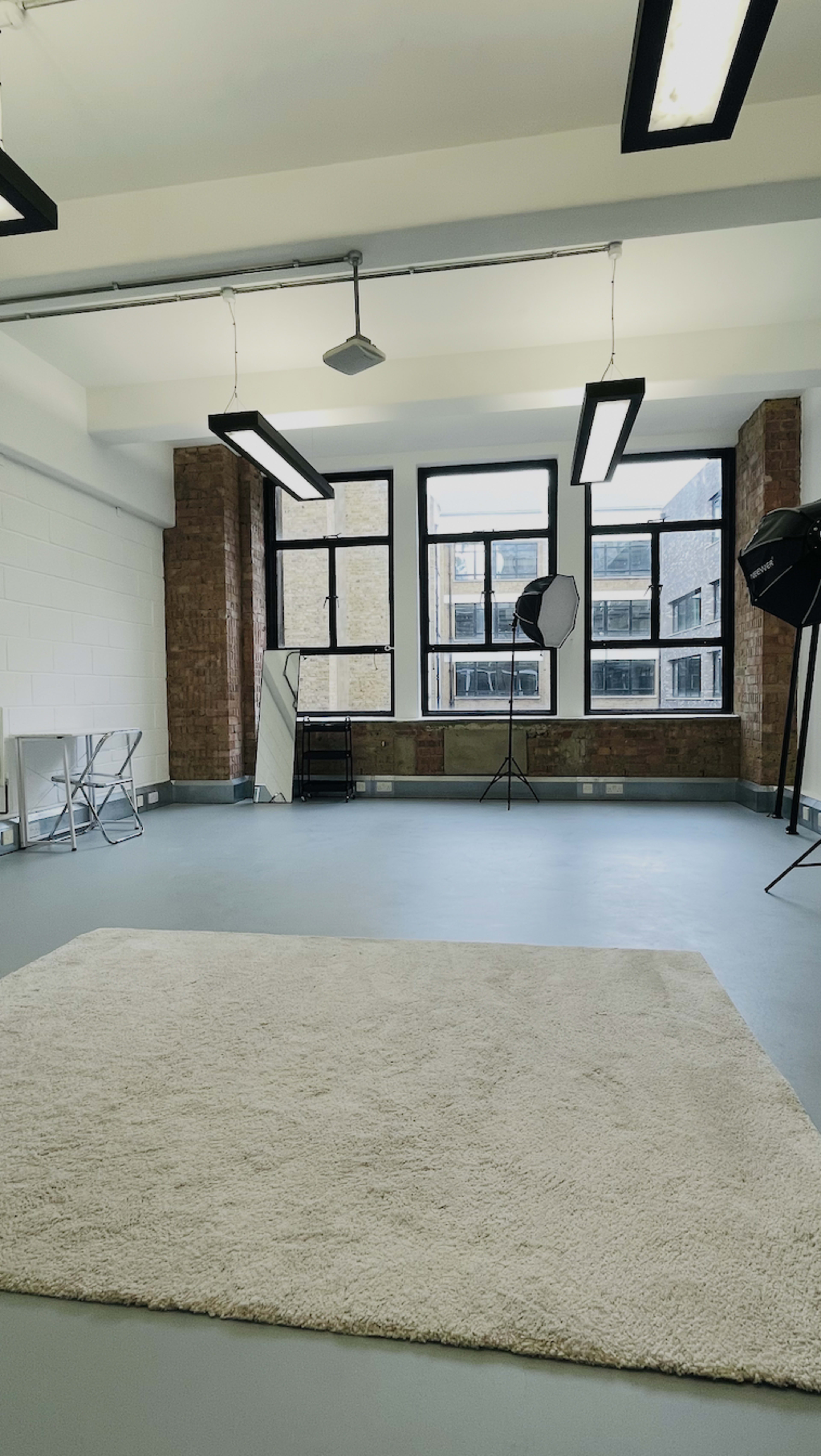 The image shows a spacious studio with a light-colored rug in the center, large windows, and photography lighting equipment.