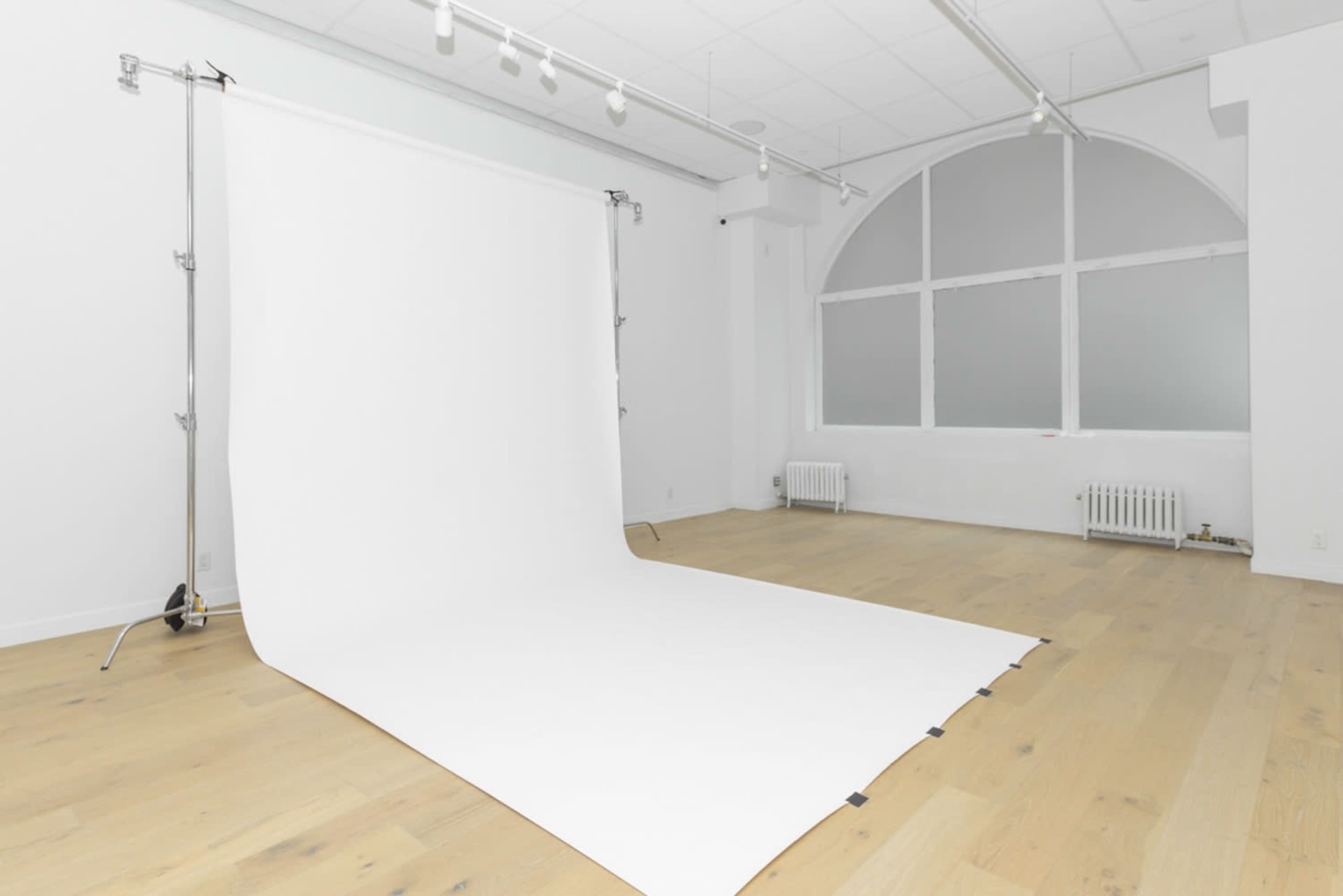 The image shows a minimalist photography studio featuring a white backdrop and wooden flooring, with large windows allowing natural light.