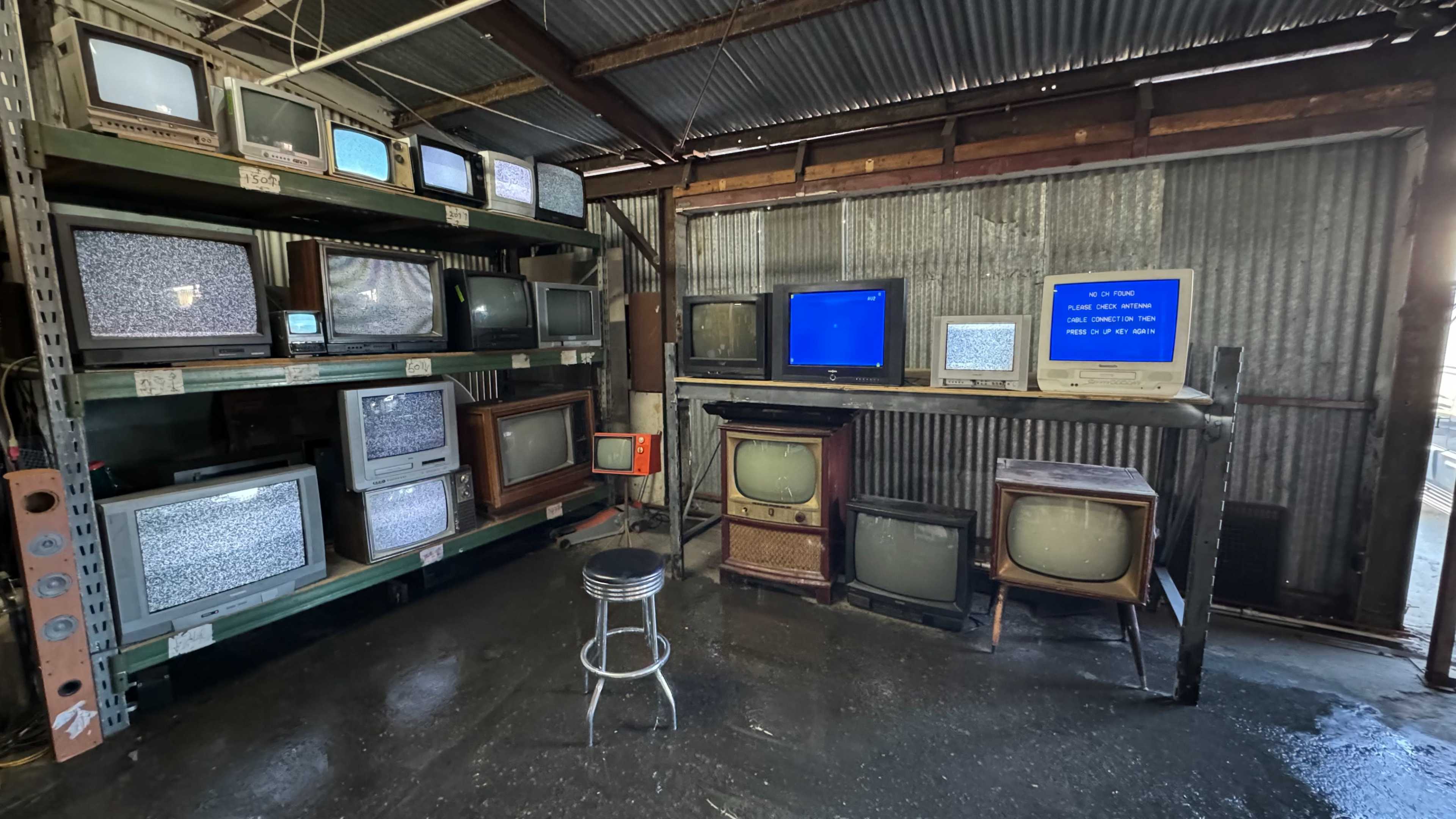 A collection of old television sets arranged on shelves and a table in a dimly lit room with a metal roof.