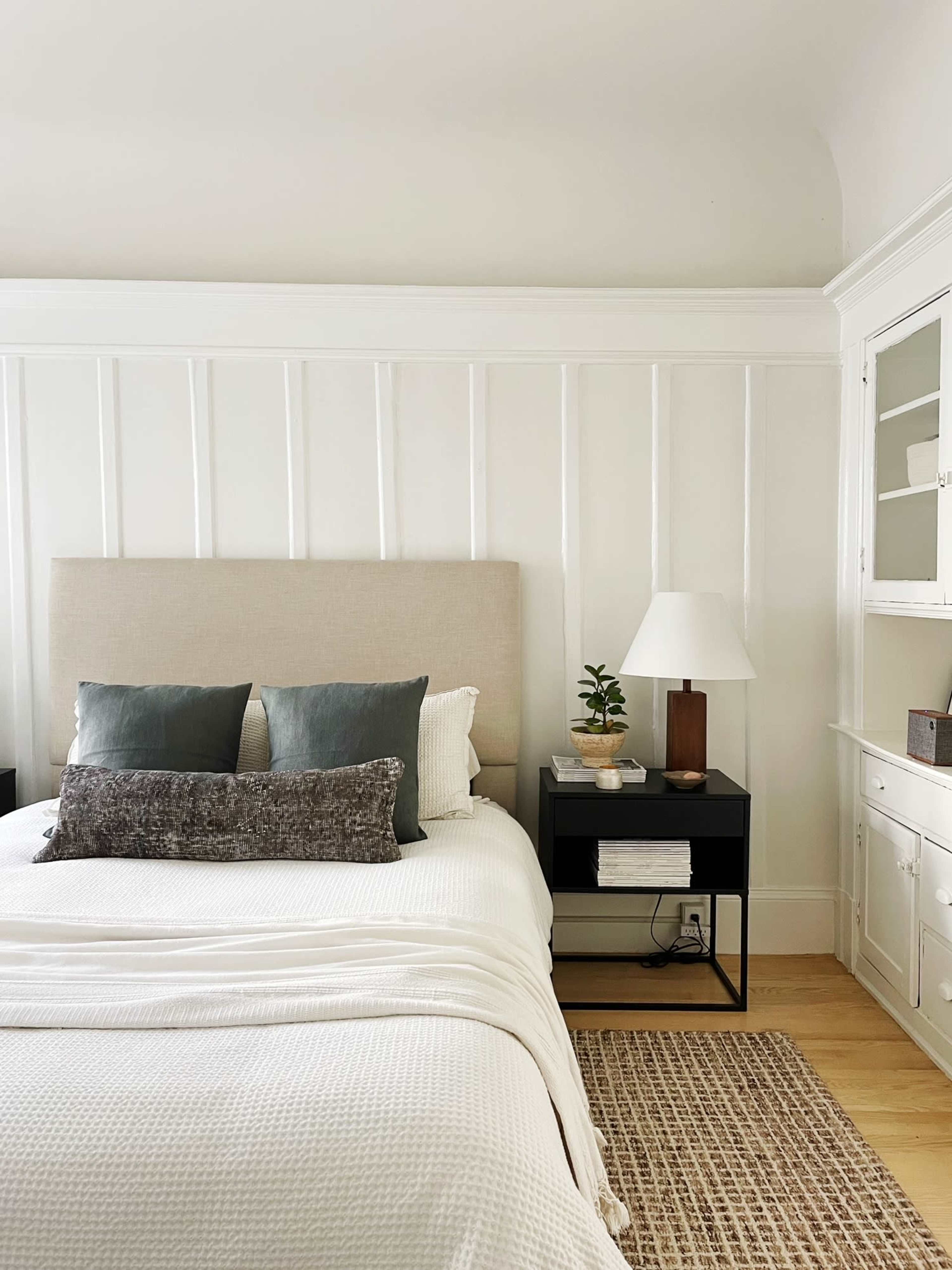 A neatly made bed with a light-colored quilt, decorative pillows, a nightstand with a lamp and a small plant, and a white paneled wall in a bright bedroom.