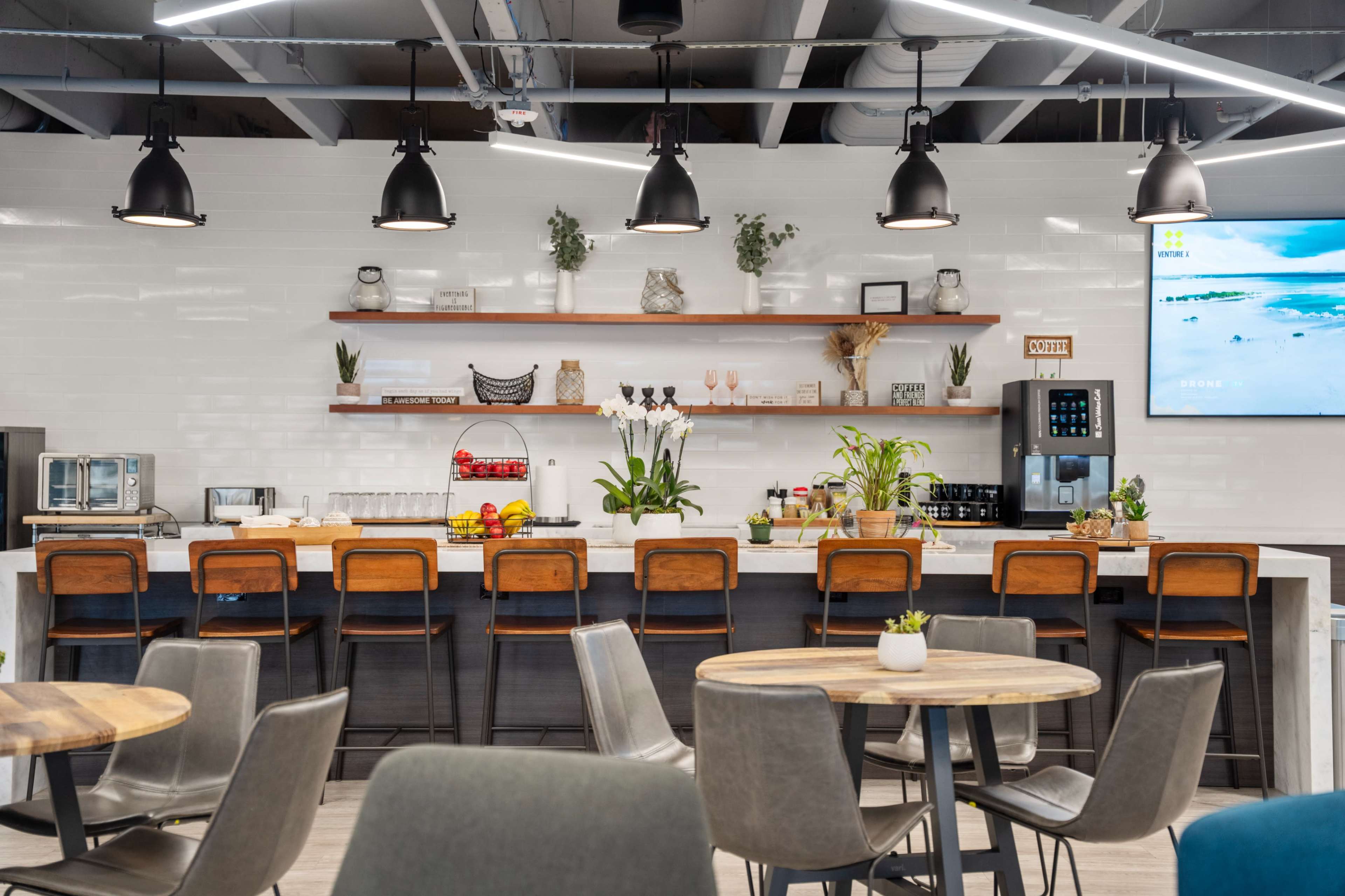 The image shows a modern kitchen area with a long bar featuring wooden stools, shelves displaying decorative items, and a variety of food and beverage stations.