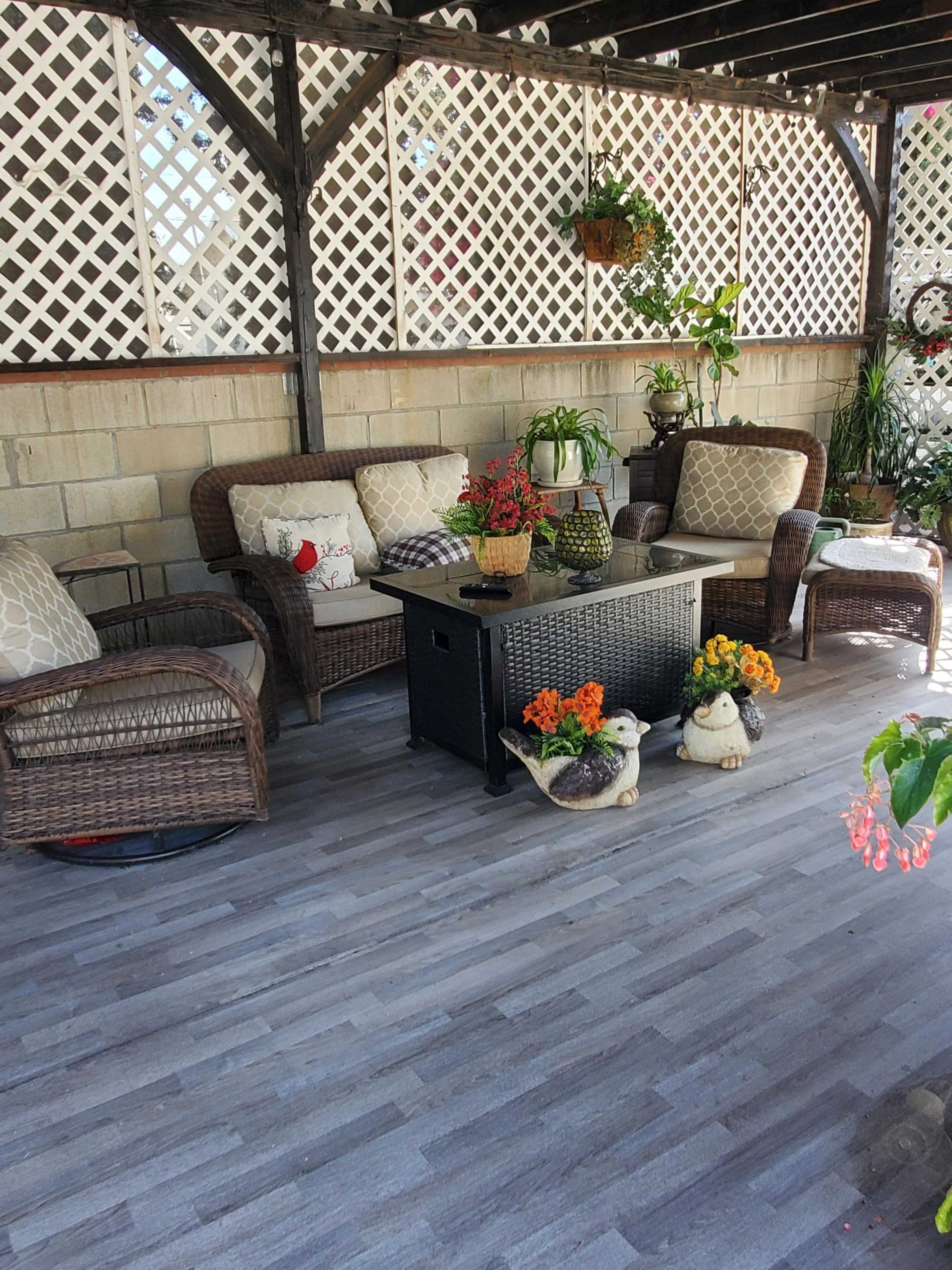 The image shows a patio area with wicker furniture, a central black table, and various potted plants and decorative items.