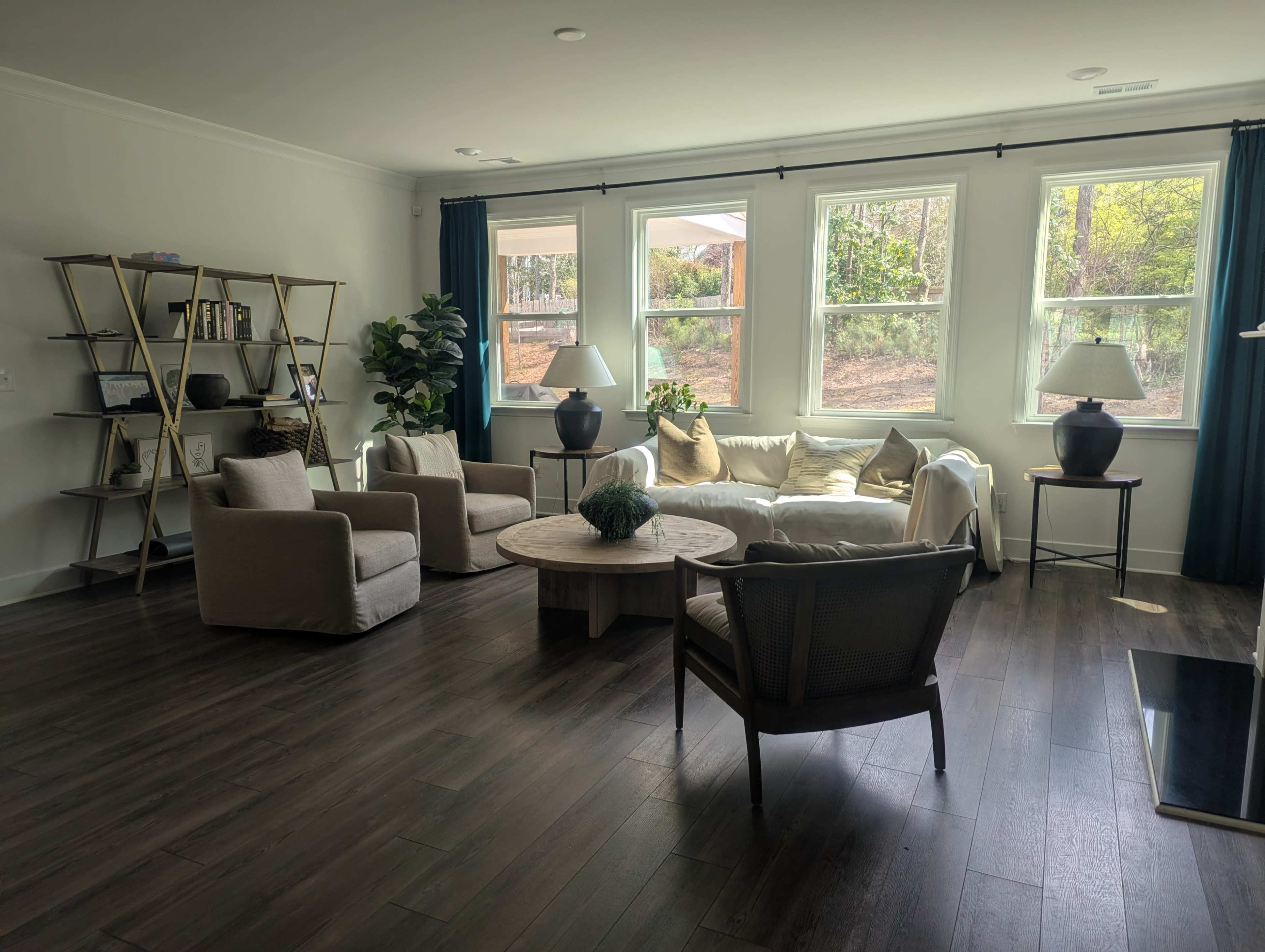 The image shows a modern living room with a circular coffee table, two armchairs, a white sectional sofa, and large windows dressed with green curtains.