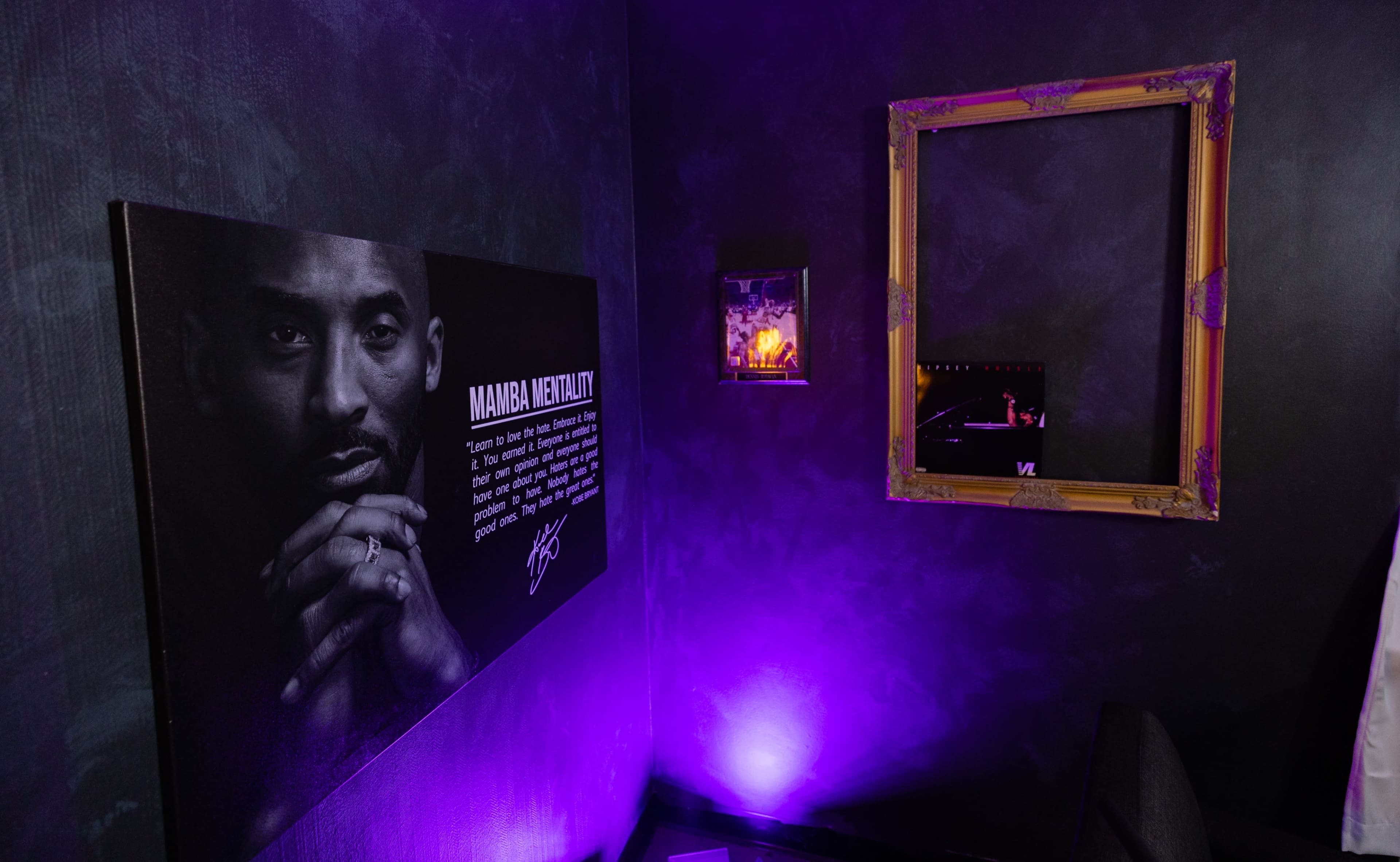 The image shows a dimly lit corner of a room featuring a large canvas of a person with a quote about "Mamba Mentality" on the wall, accompanied by a framed mirror and purple lighting.
