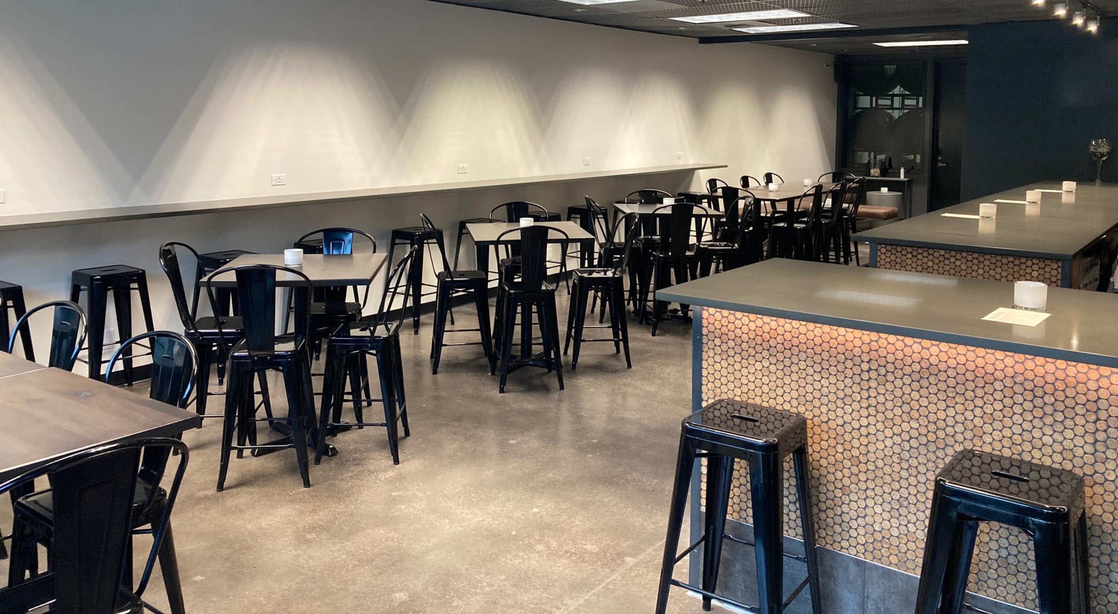 The image shows a spacious, modern interior of a café or restaurant featuring black metal stools and tables on a polished concrete floor.
