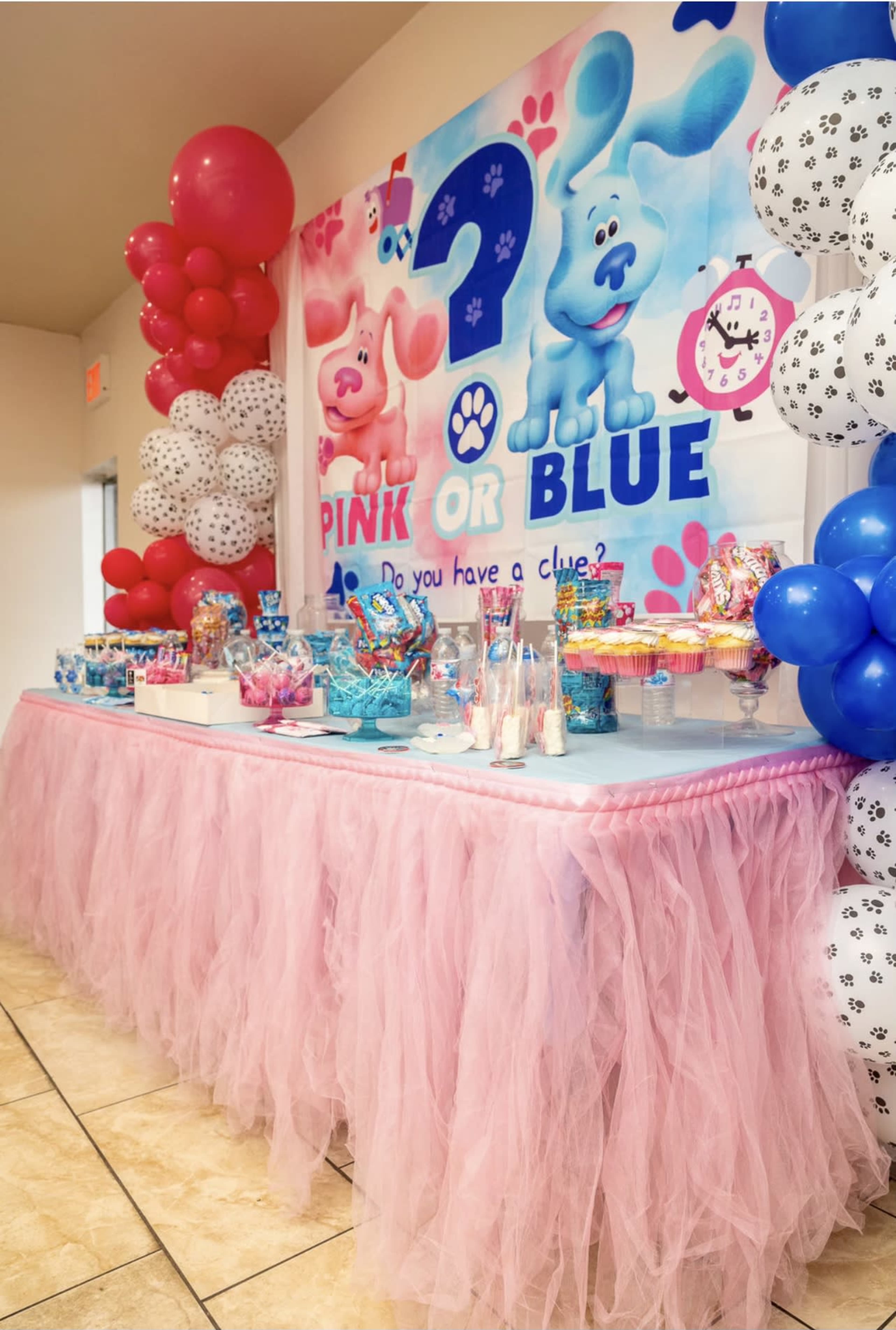 A decorated table with pink tulle, surrounded by balloons, features a colorful dessert spread for a "Pink or Blue" themed party.