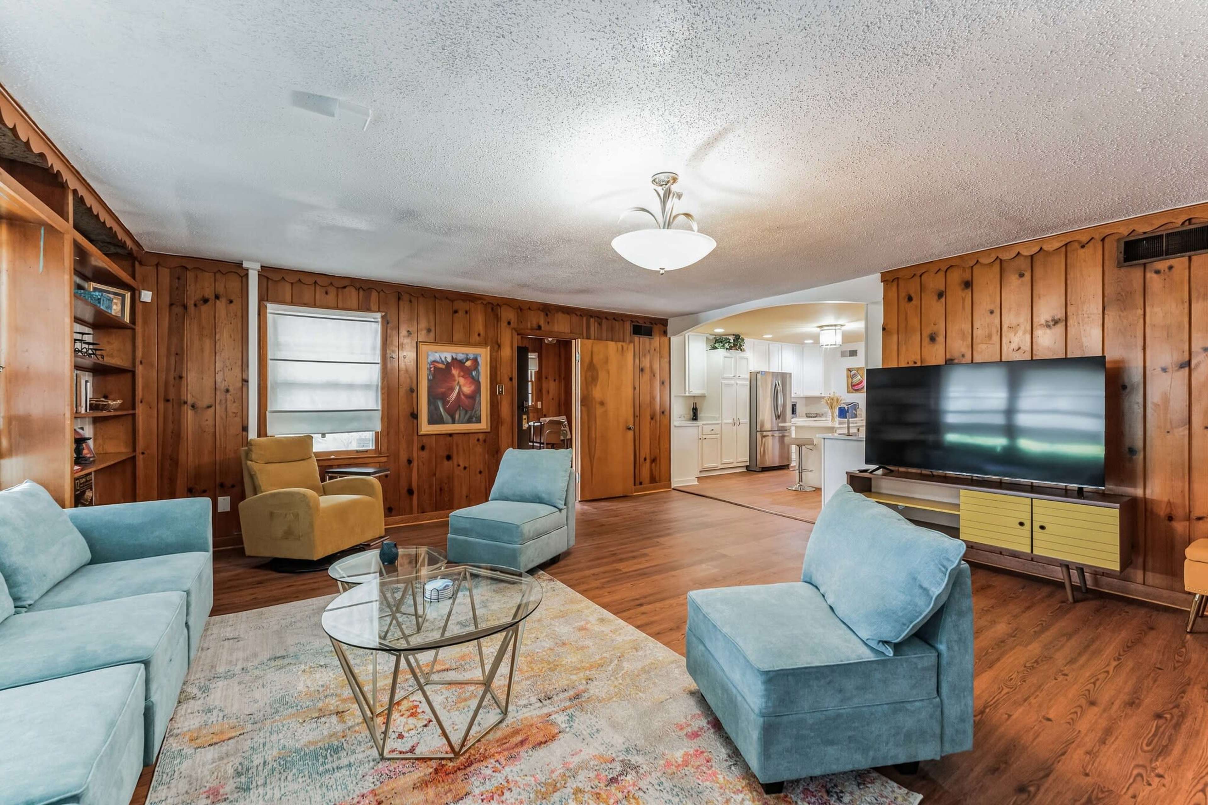 A living room with wooden-paneled walls, a glass coffee table, teal armchairs, and a large TV mounted on a media console.