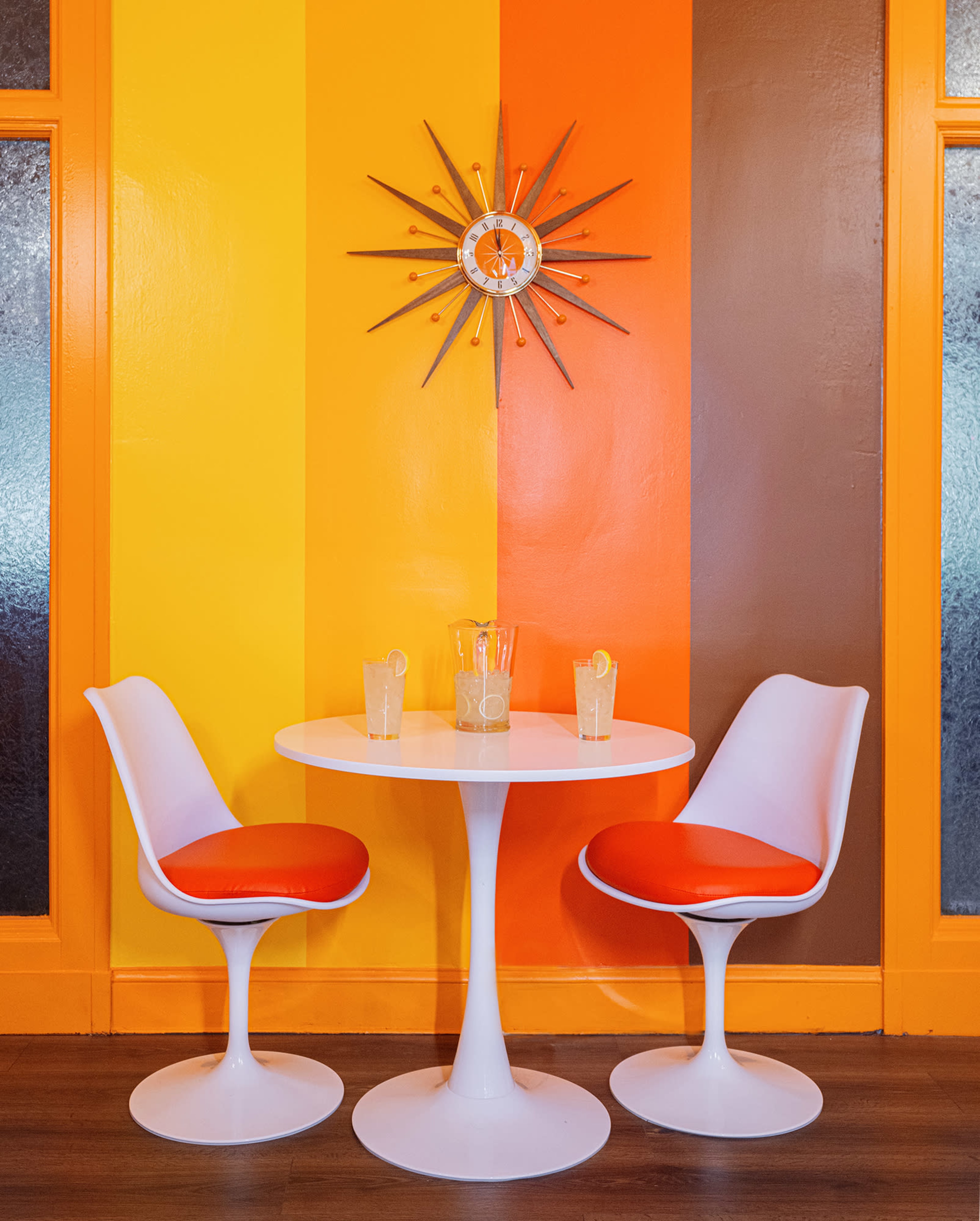 A round white table with two orange-accented chairs is set against a striped wall of yellow, orange, and brown, with a starburst wall clock above.