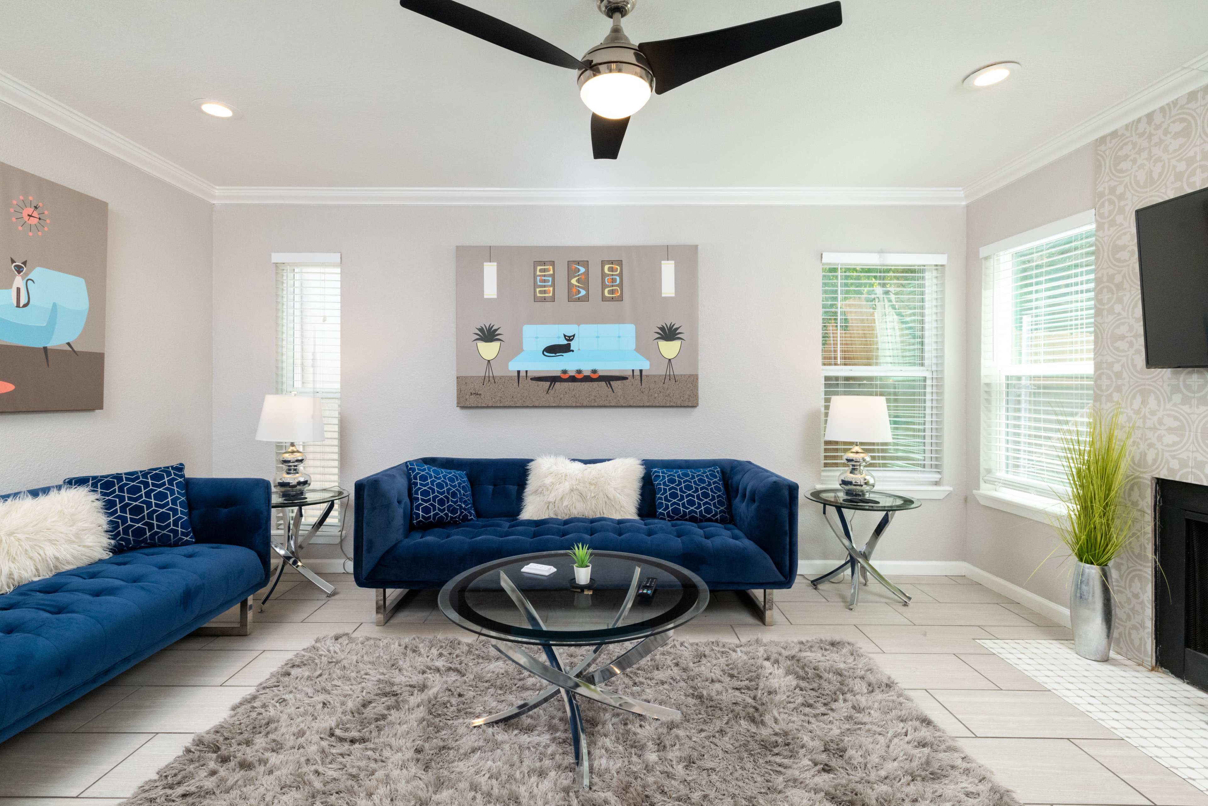 The image shows a modern living room with blue couches, a round glass coffee table, and decorative art on the walls.