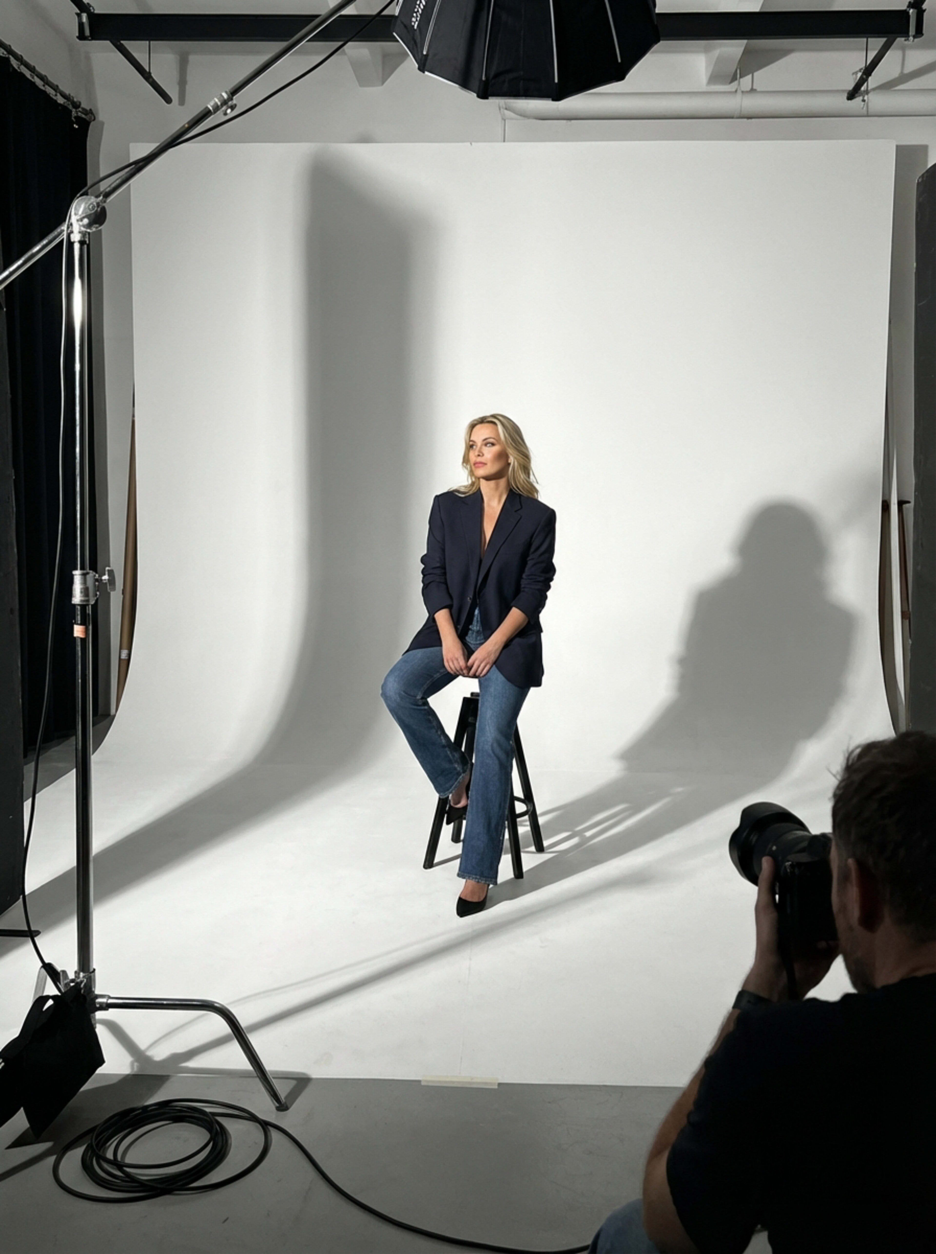 A person sits on a stool in a photography studio, with soft lighting and a plain backdrop.