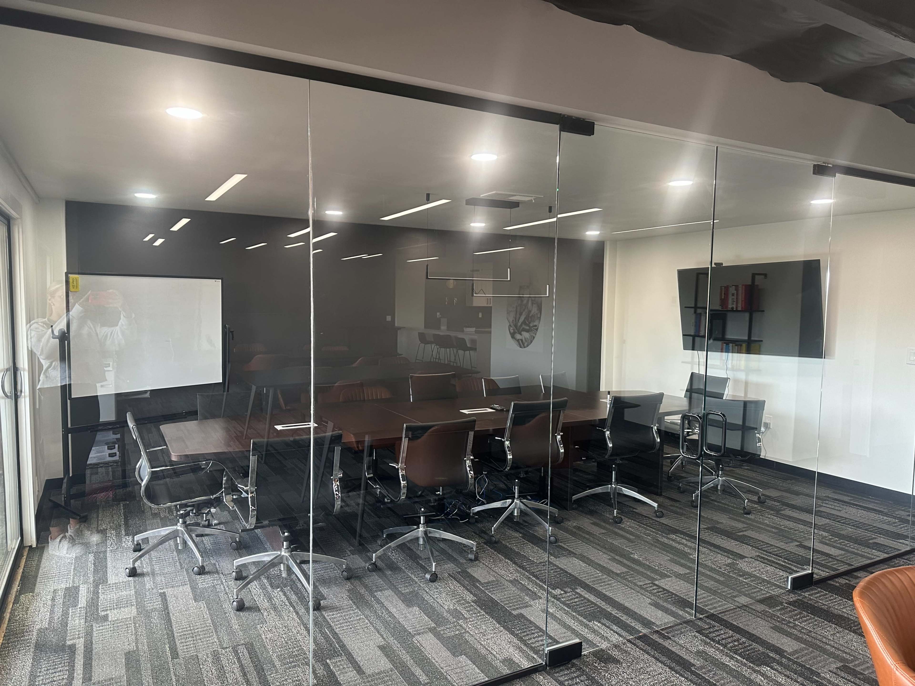 A modern conference room with a large glass wall, featuring a long table surrounded by chairs and a wall-mounted screen.