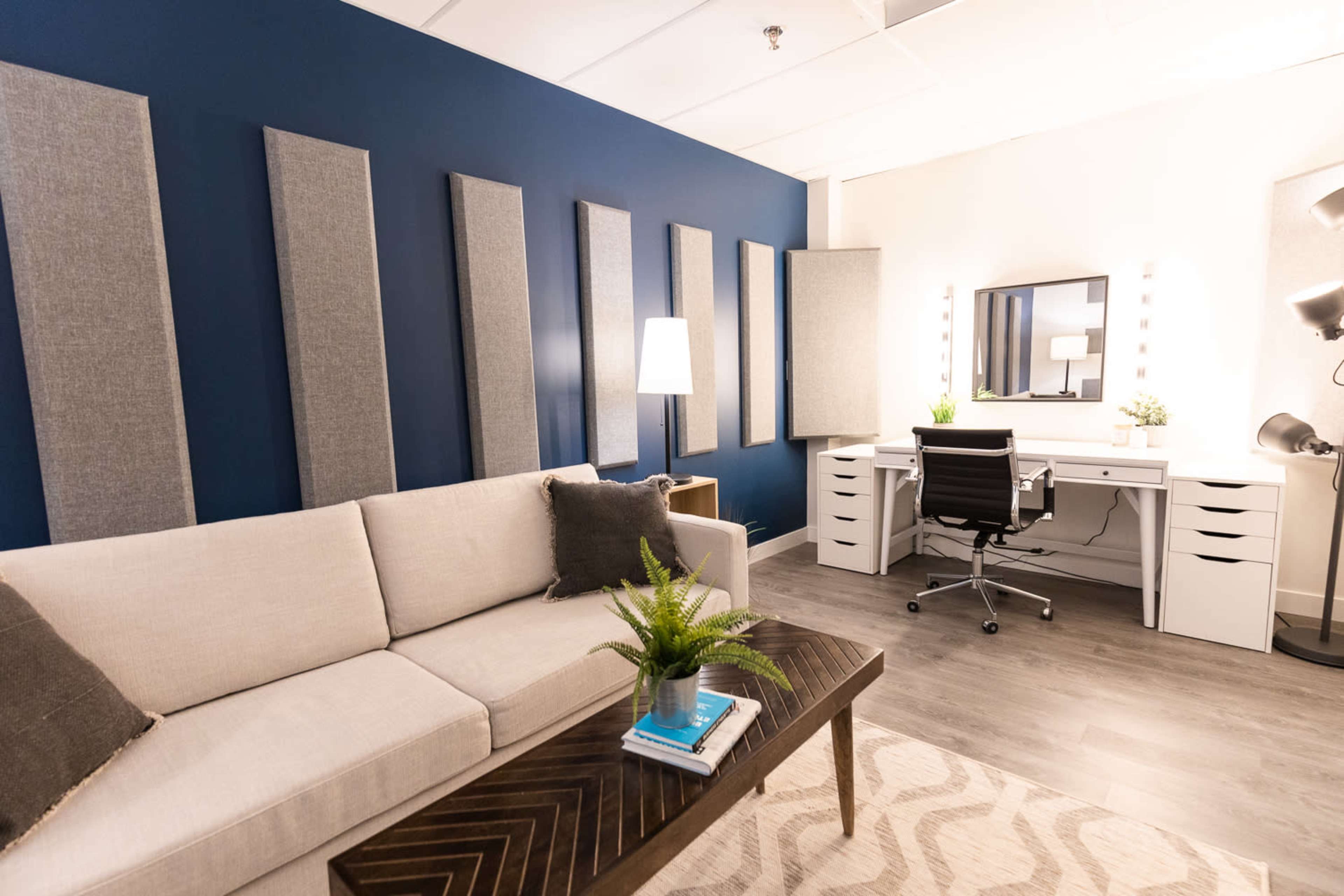 A modern office space with a light-colored sofa, a coffee table, and a white desk with a mirror, illuminated by both natural and artificial lighting.