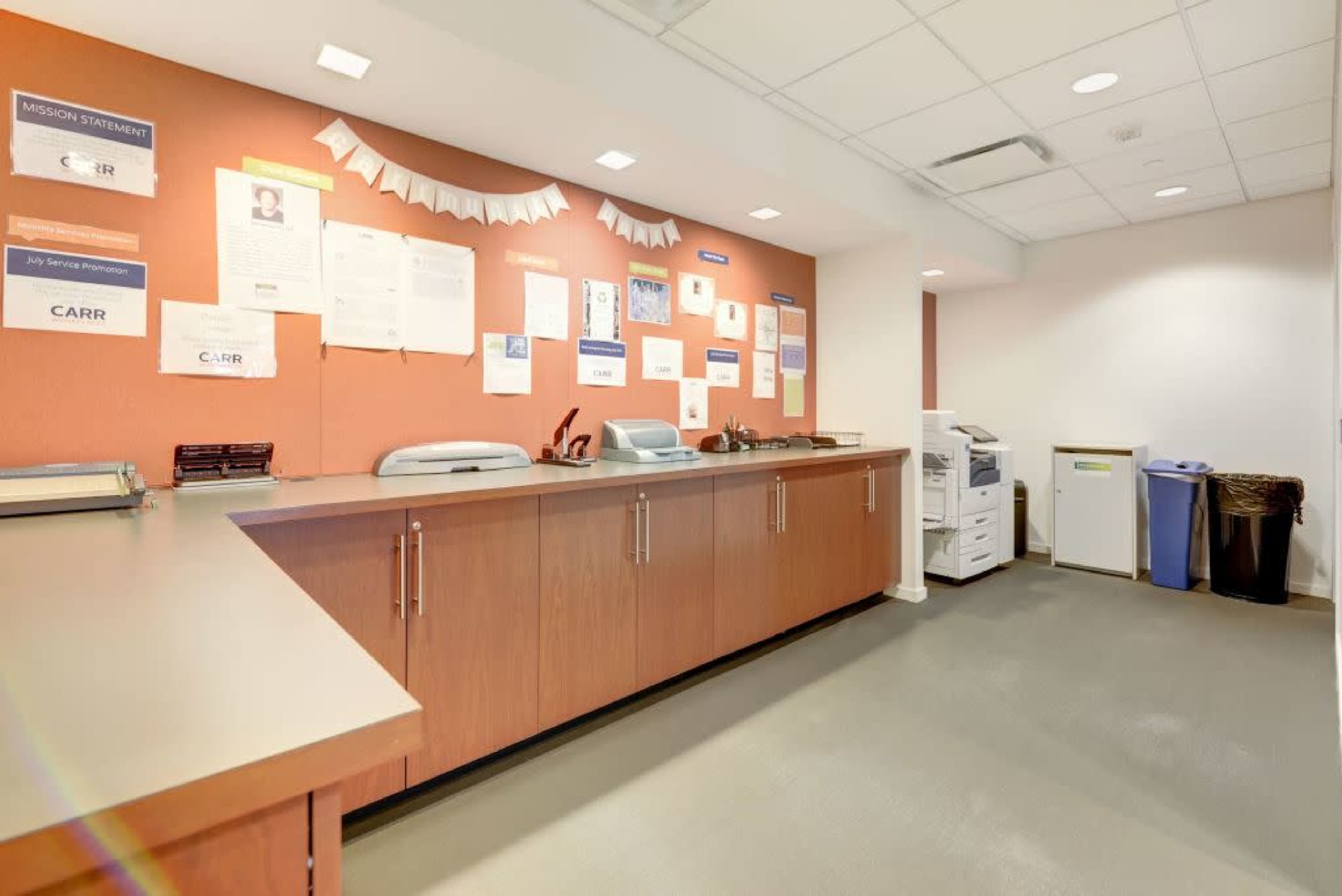 The image shows an office reception area with a wooden counter, a copier, and several informational posters on a orange wall.