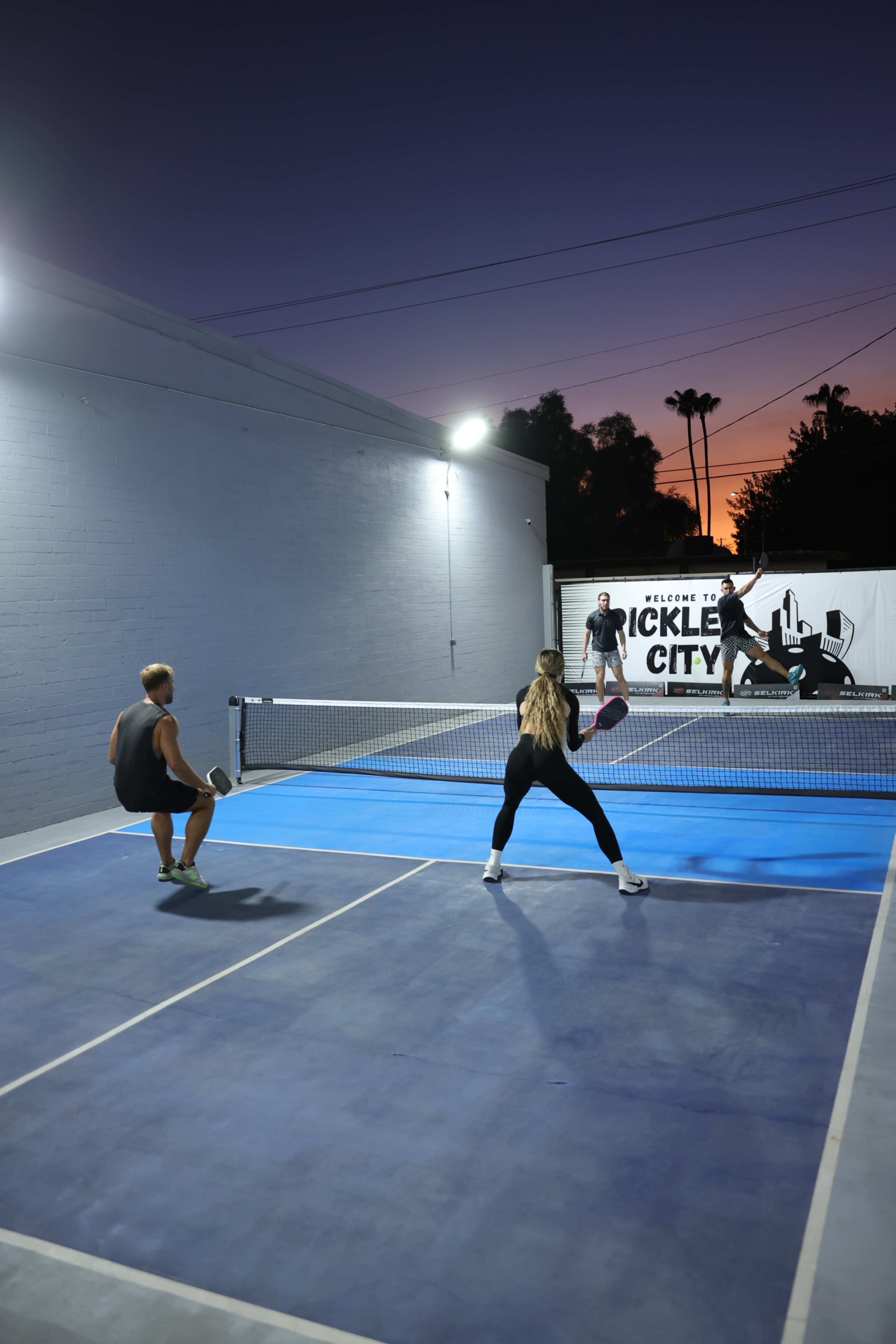 Two players are positioned on a blue paddle tennis court at dusk while a third player prepares to serve on the opposite side.