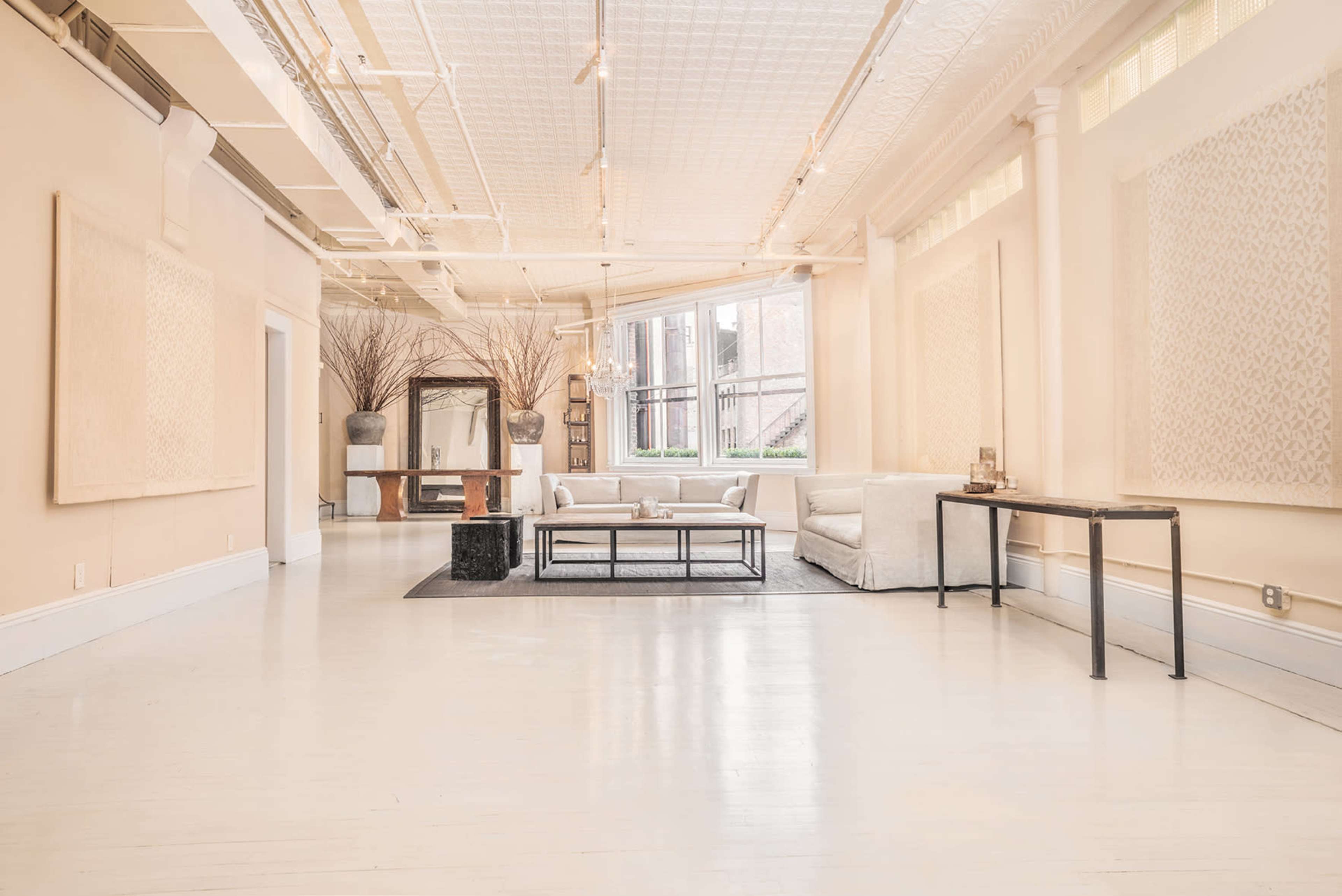 The image shows a spacious, well-lit room with light-colored walls, a seating area with a white sofa, and a coffee table, complemented by a large window and decorative wall art.