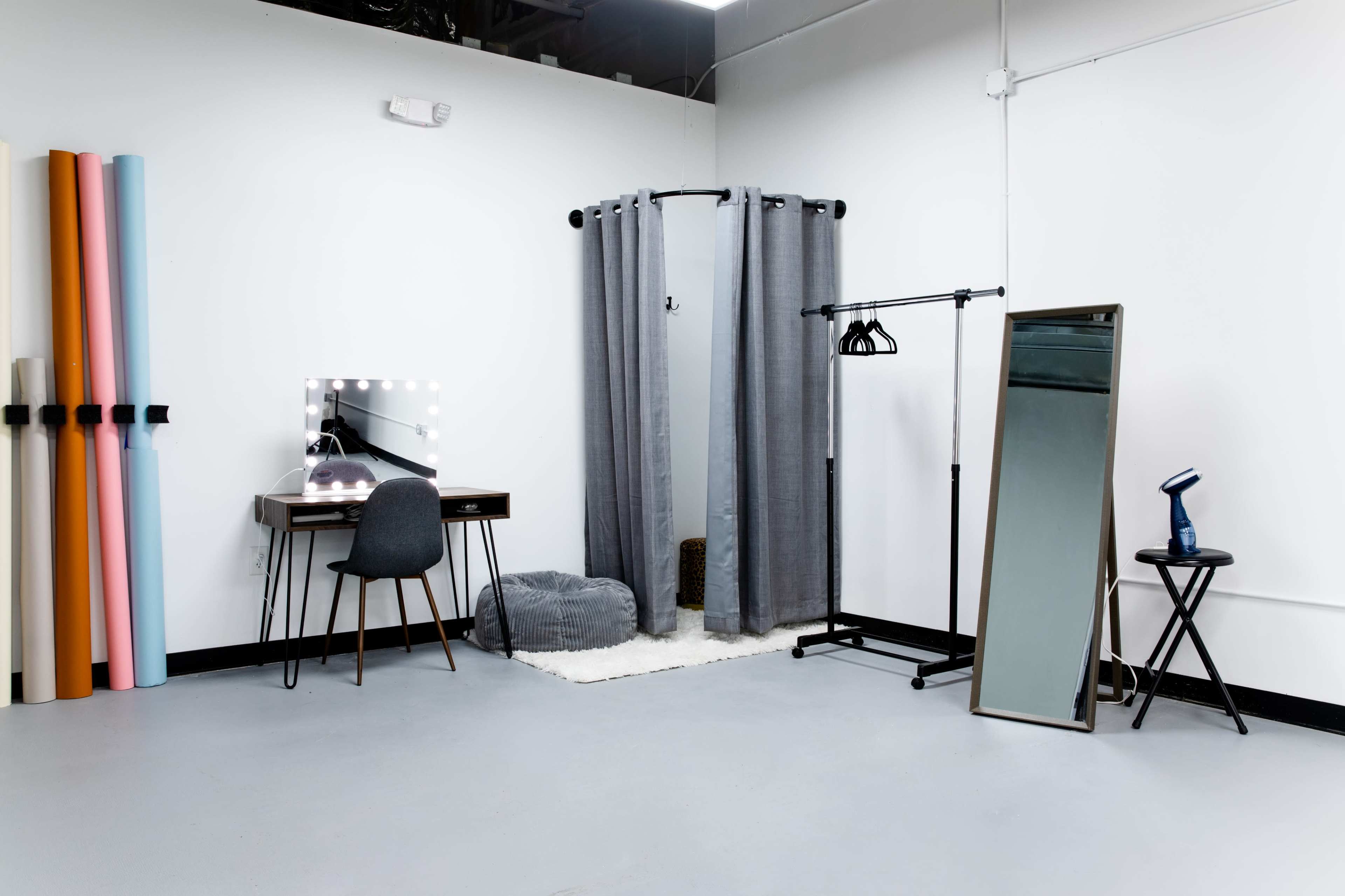The image shows a minimalist studio space featuring a dressing area with a mirror, a vanity table, a garment rack, and several rolls of colored paper against the wall.