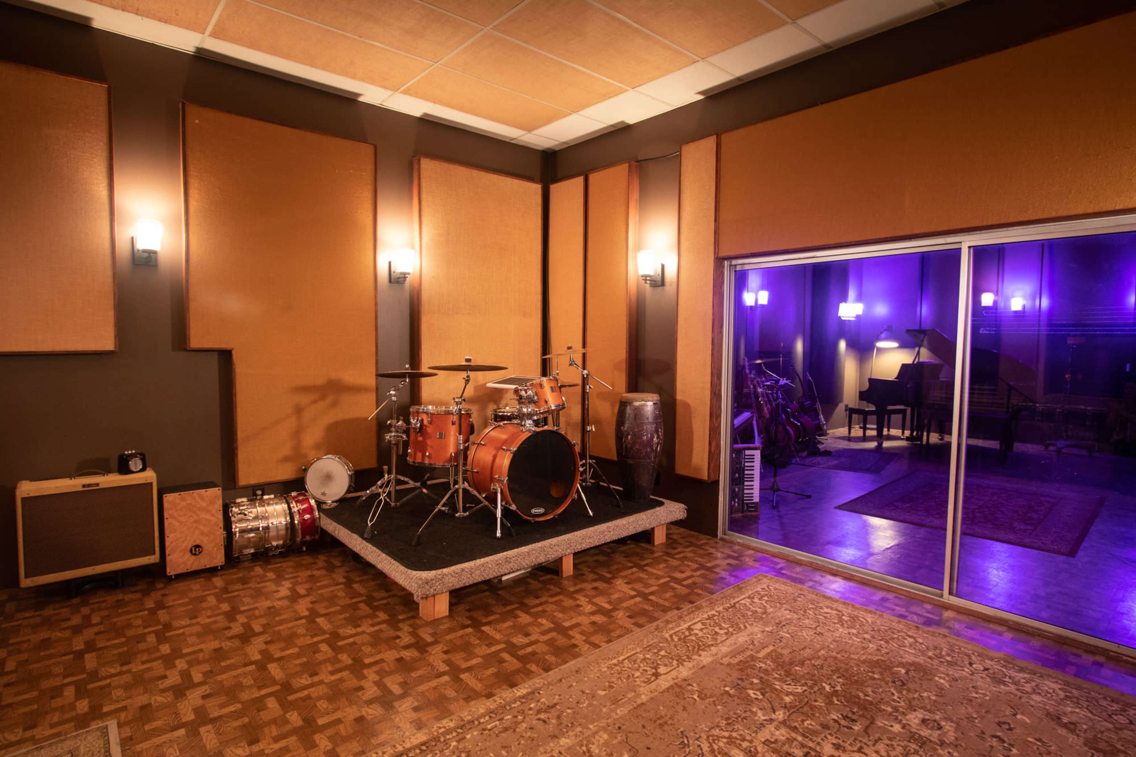 A music studio features a drum set on a raised platform, surrounded by soundproof walls and visible musical instruments through a glass sliding door.