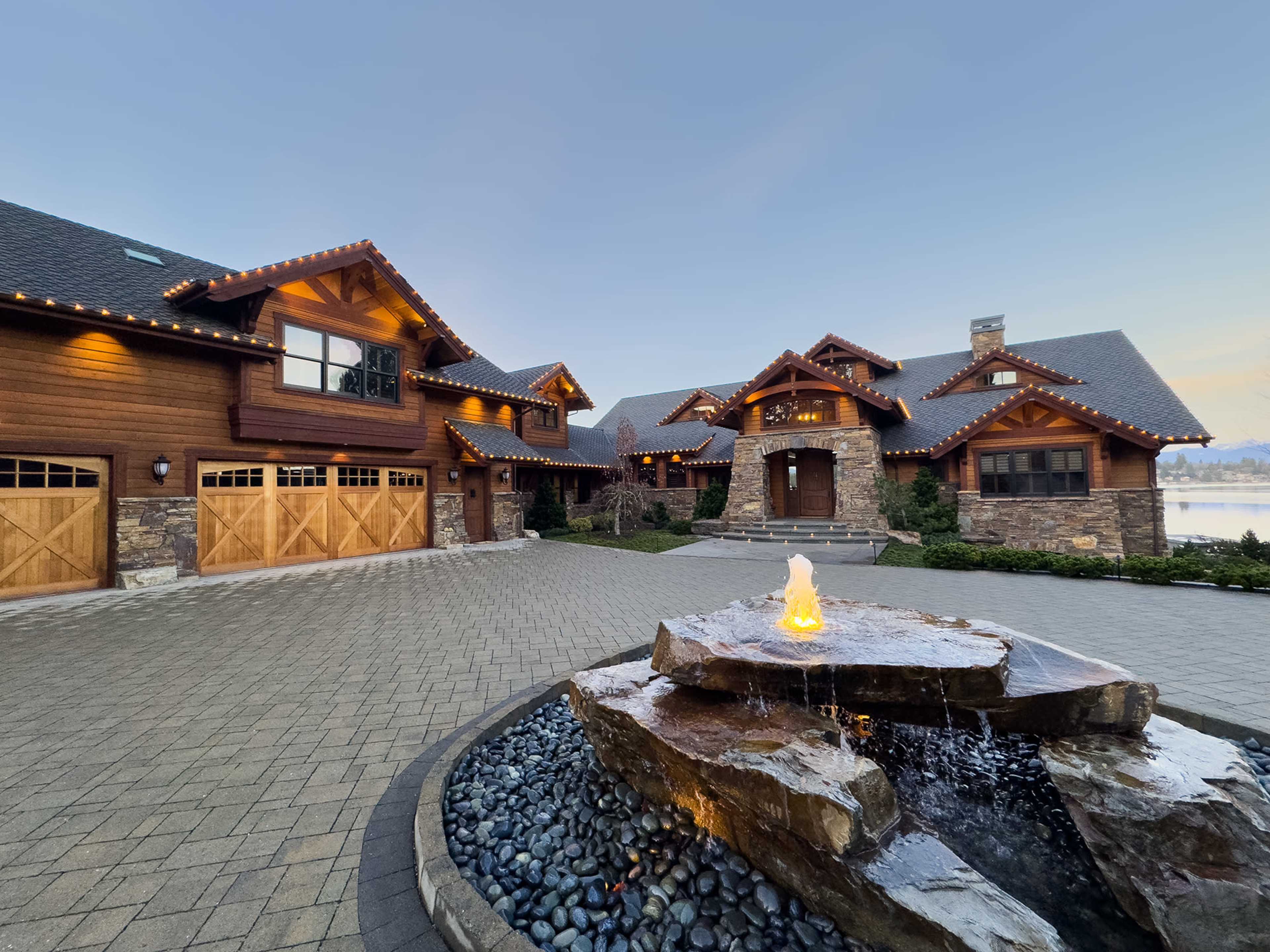 A large, rustic-style house with stone accents and a circular driveway features a water fountain made of rock and pebbles in the foreground.