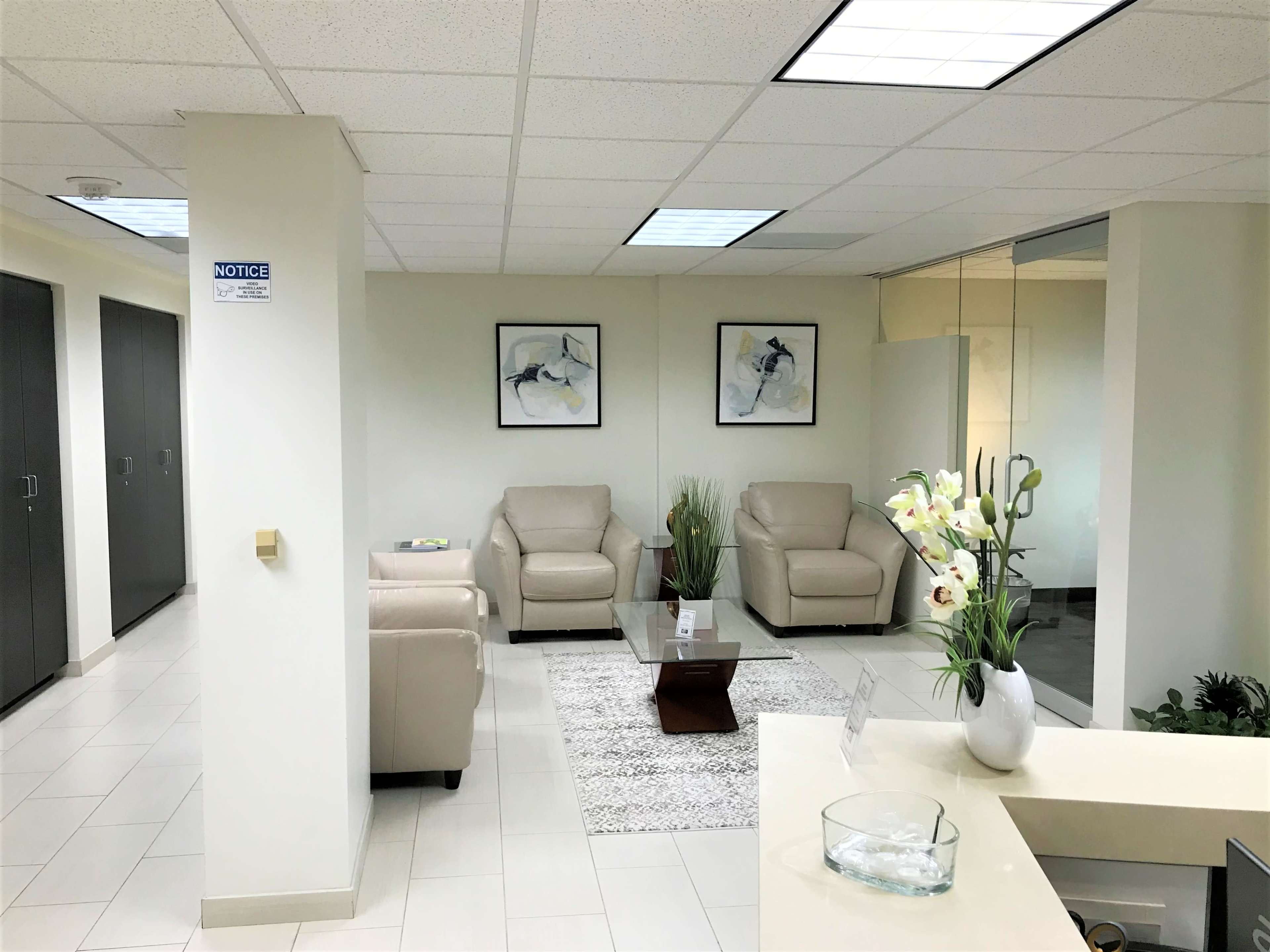 A modern waiting area featuring two light-colored chairs, a glass coffee table on a rug, and decorative plants, with framed artwork on the walls.