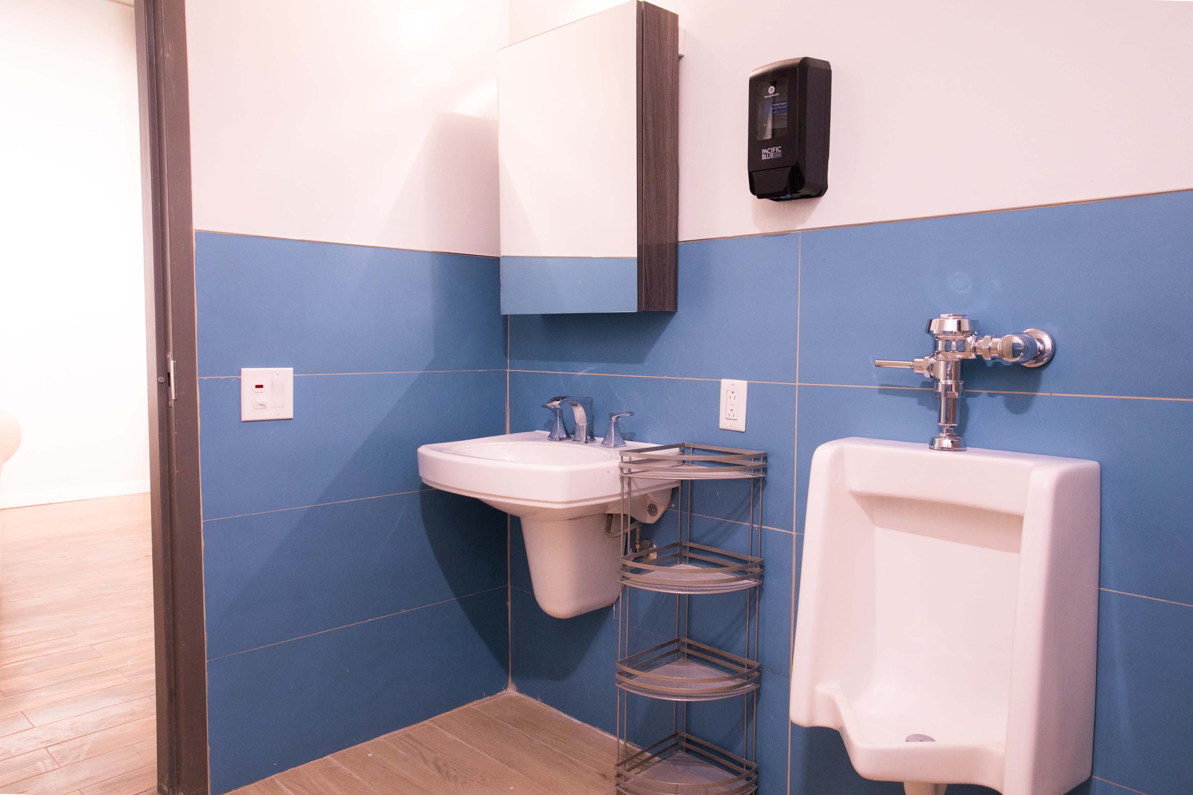 The image shows a bathroom with a sink, a wall-mounted urinal, a mirror, and a soap dispenser on a blue-tiled wall.