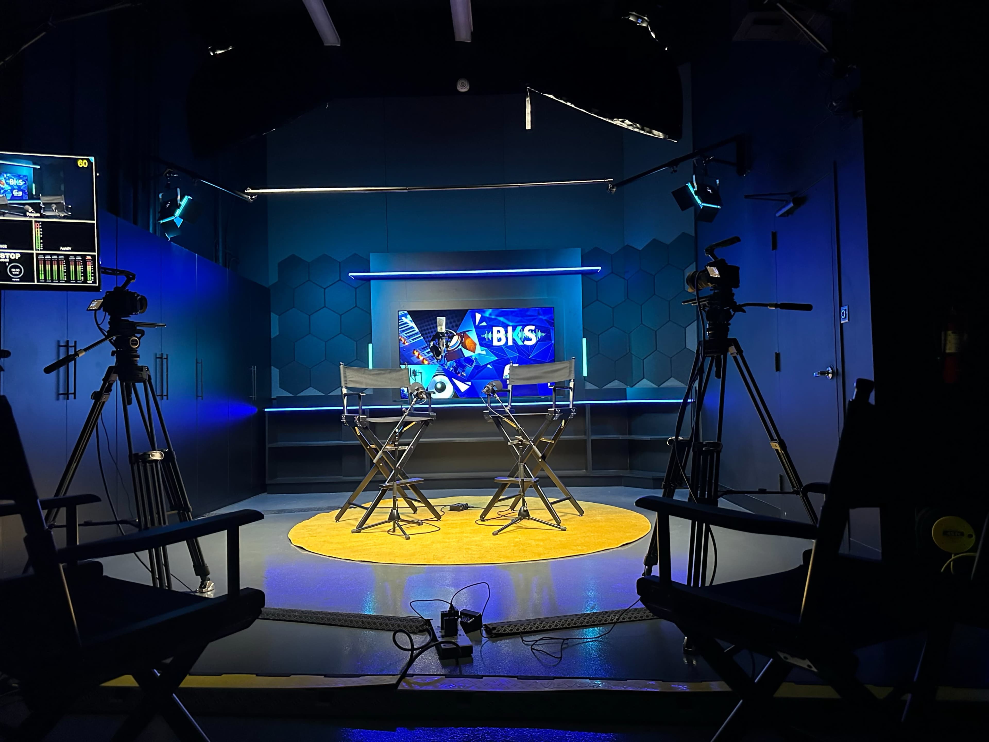 A television studio setup with two director's chairs facing a brightly lit screen, flanked by cameras and staging equipment.