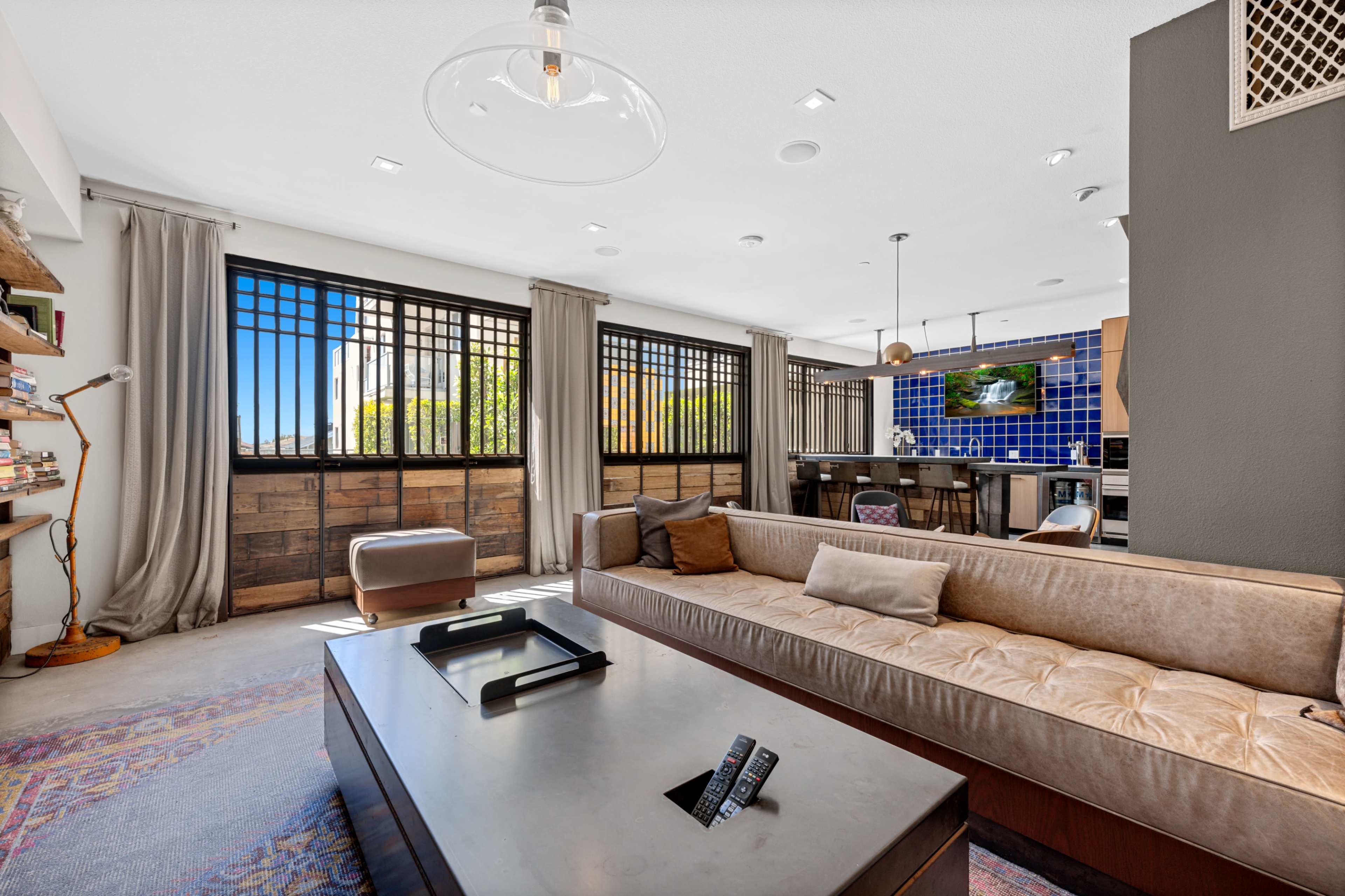 The image shows a modern living room with large windows, a brown sofa, a coffee table, and a kitchen area in the background.