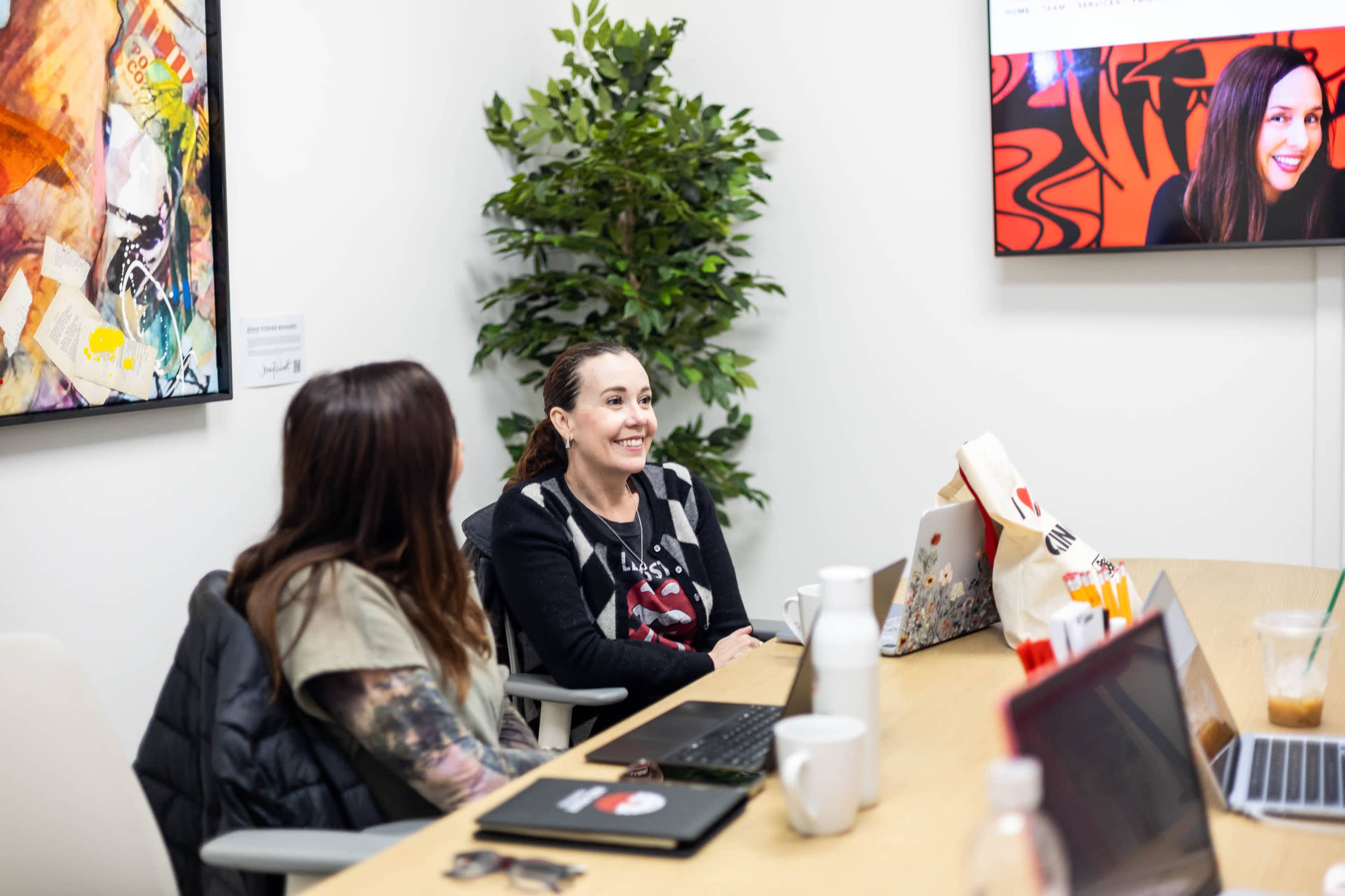 Two women sit at a conference table with laptops and drinks, while a third woman appears on a wall-mounted screen.