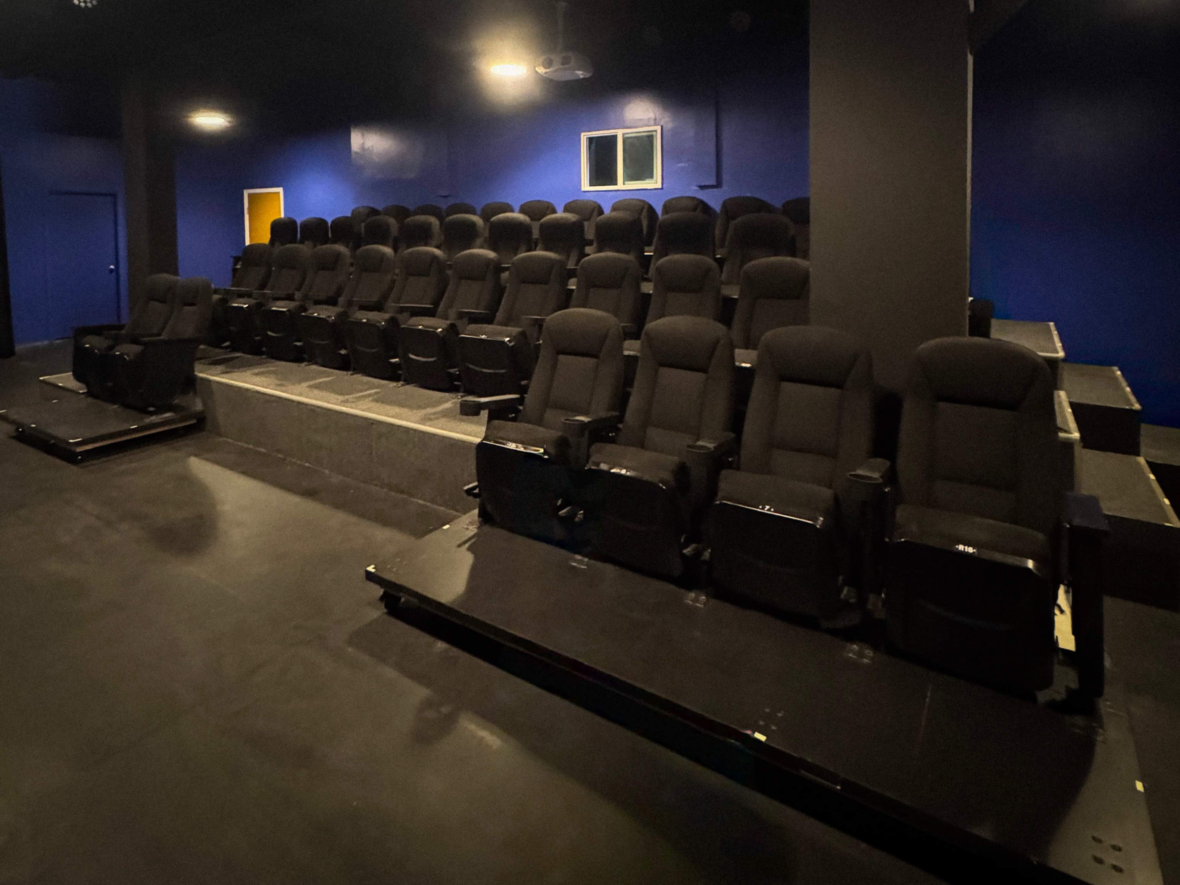 The image shows a dark, empty theater with rows of black reclining seats and a blue wall in the background.