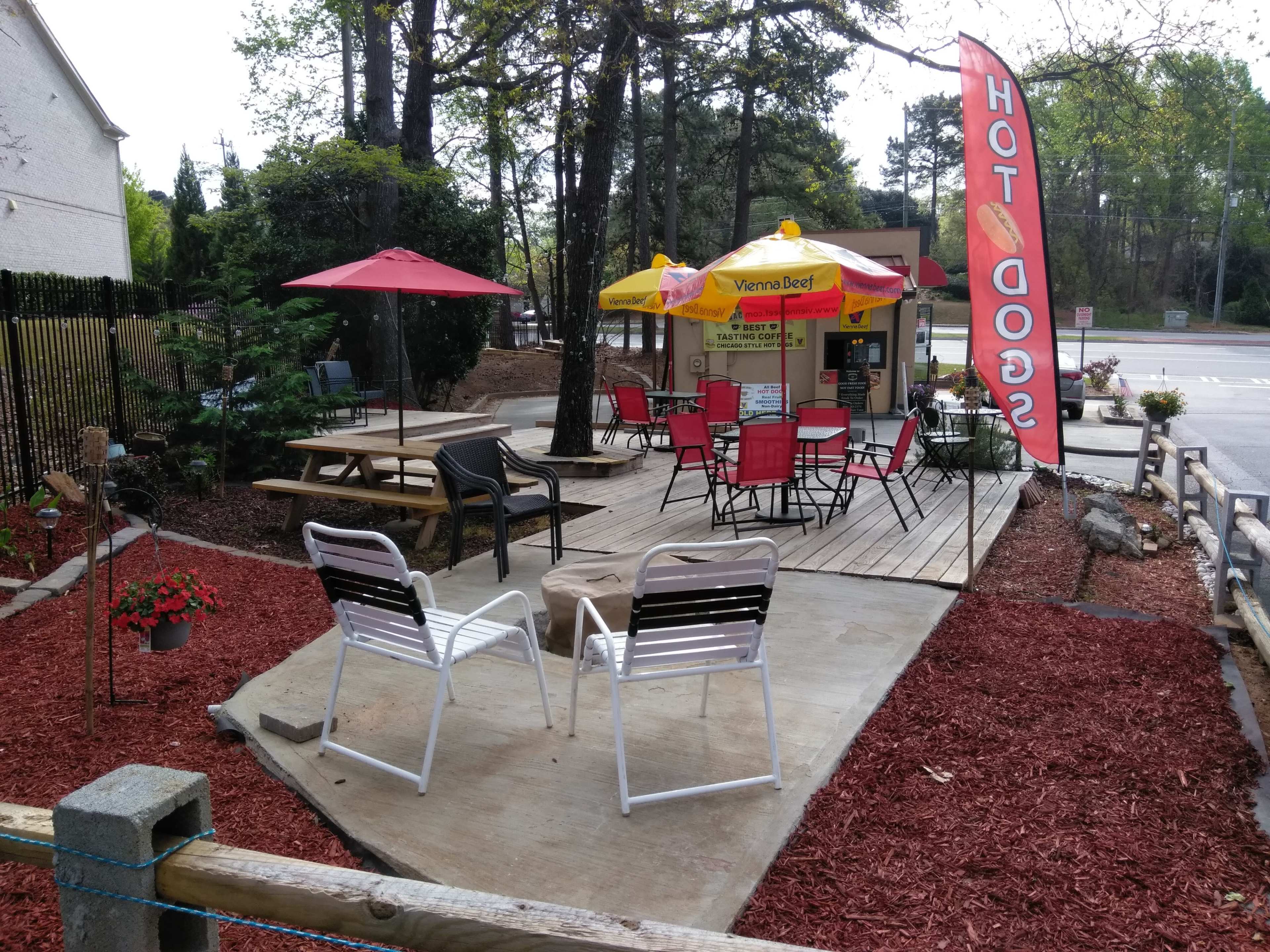 A small outdoor seating area with tables and chairs is set up next to a hot dog stand, surrounded by flower beds and a red mulch landscape.