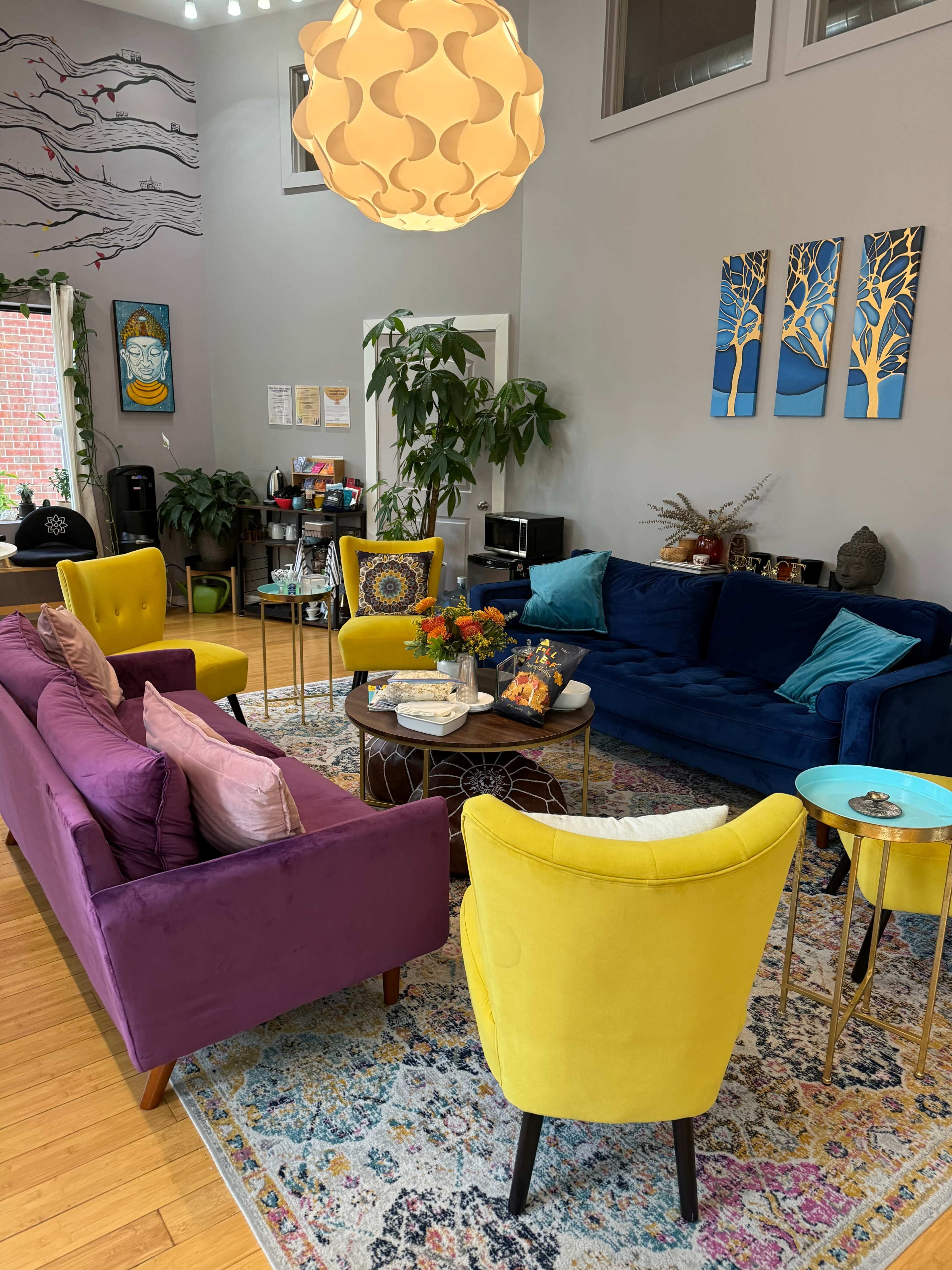 The image depicts a cozy living room with colorful furniture, including a purple sofa, teal sofa, and yellow chairs, arranged around a circular coffee table on a patterned rug.
