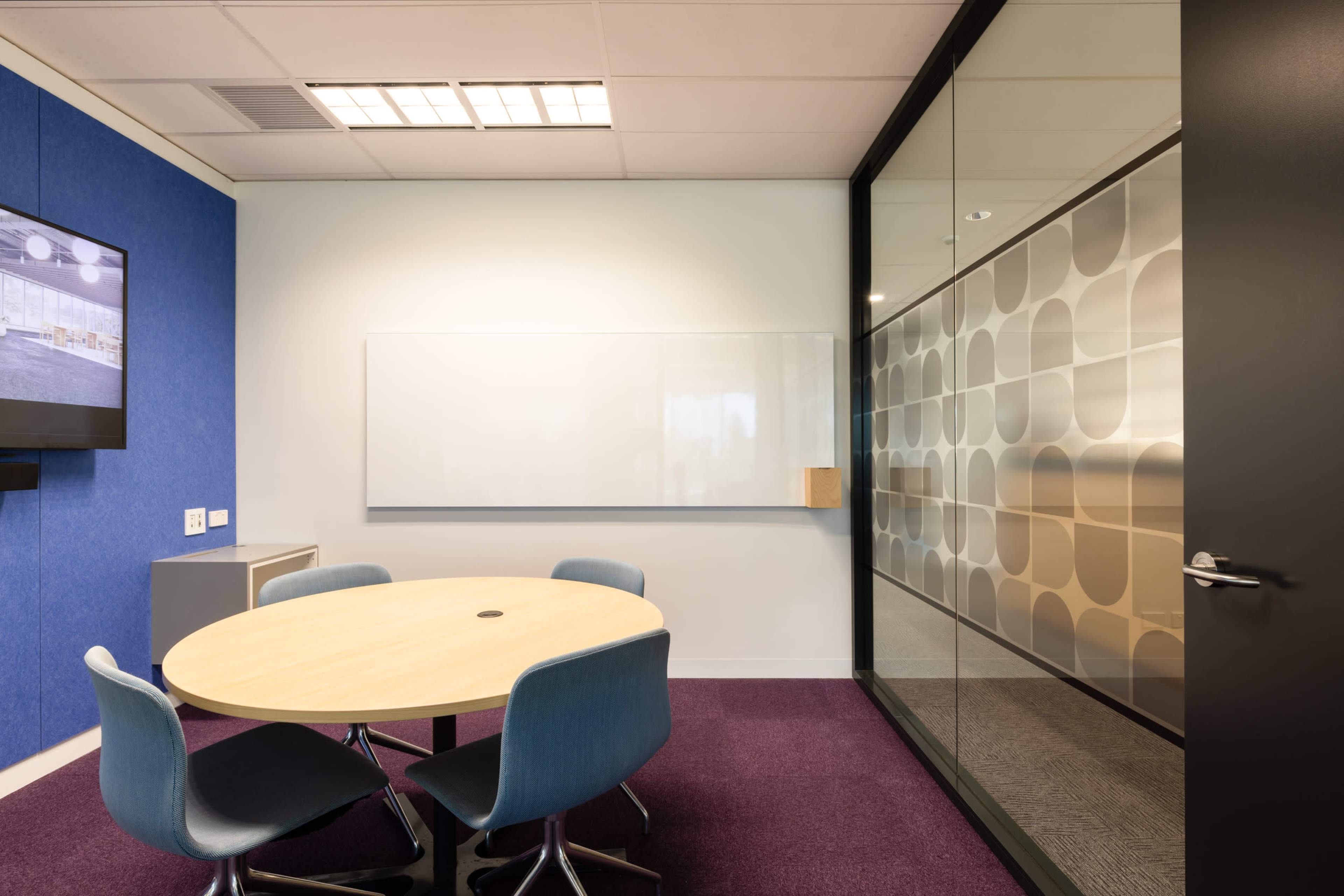 A small meeting room features a round table with chairs, a whiteboard, and a glass wall displaying a patterned design.