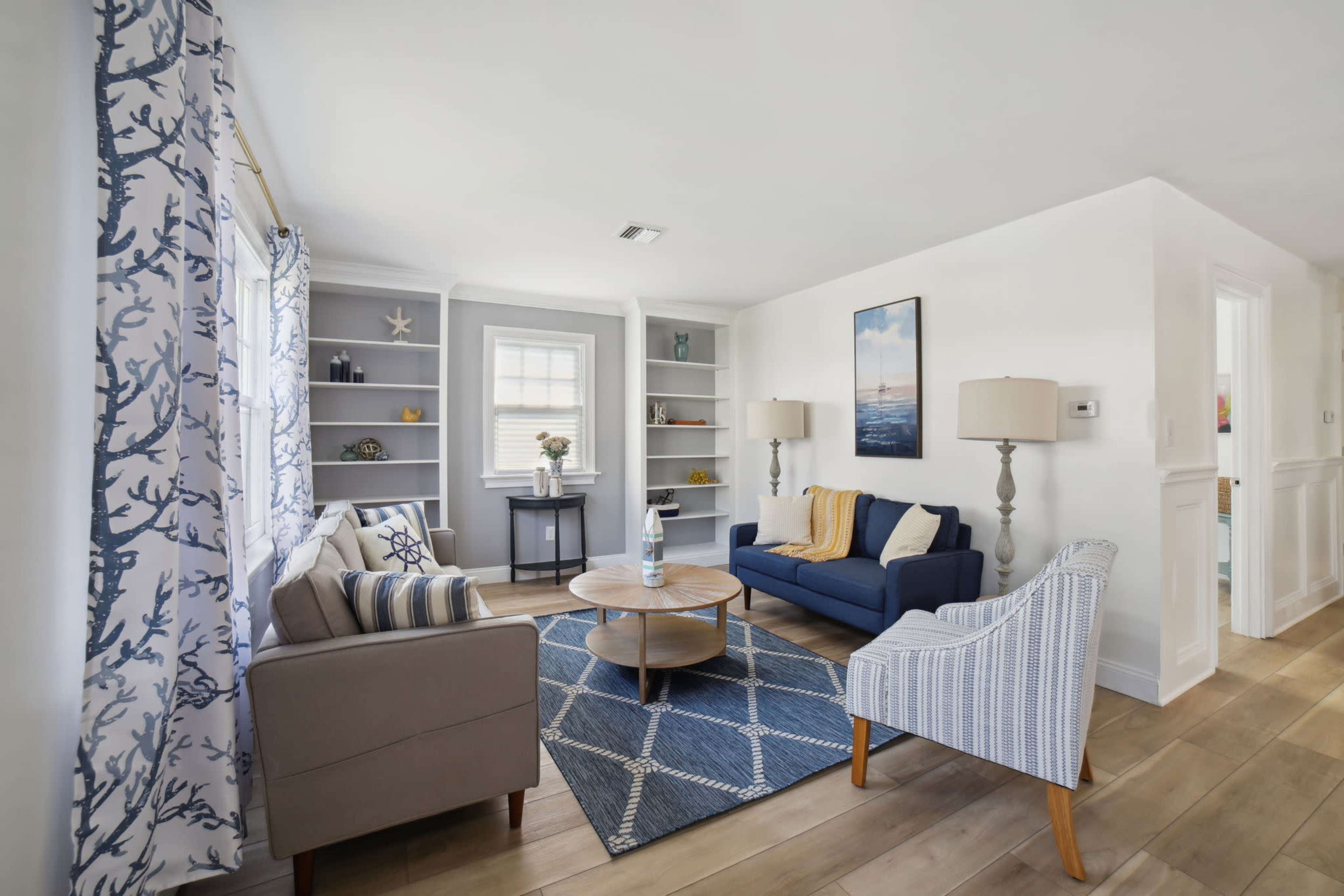 A modern living room with a blue sofa, an armchair, a round coffee table, decorative shelves, and large windows with patterned curtains.