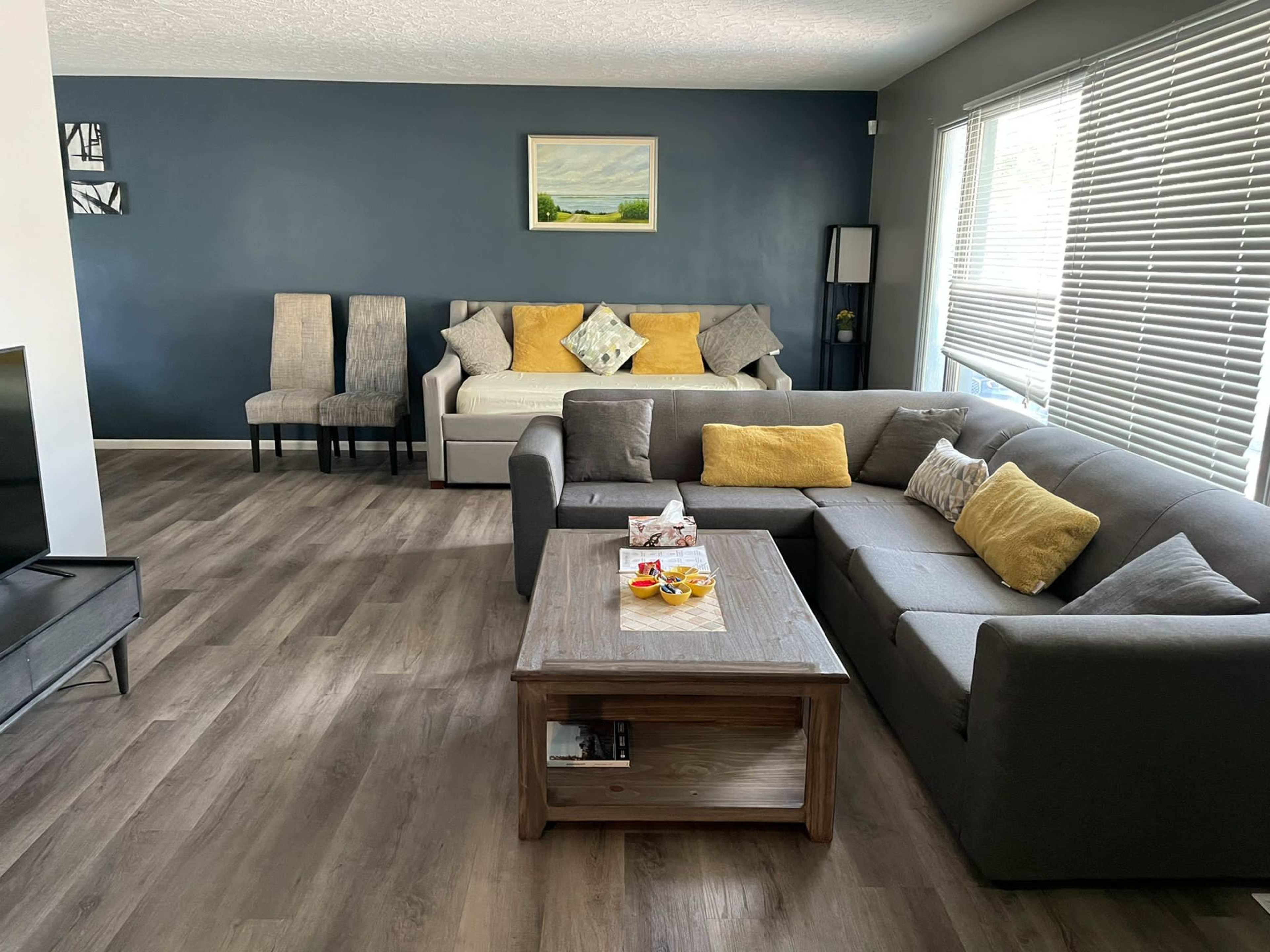 The image shows a modern living room with a gray sectional sofa, a wooden coffee table, a light-colored daybed, and two upright chairs against a blue wall, along with large windows providing natural light.