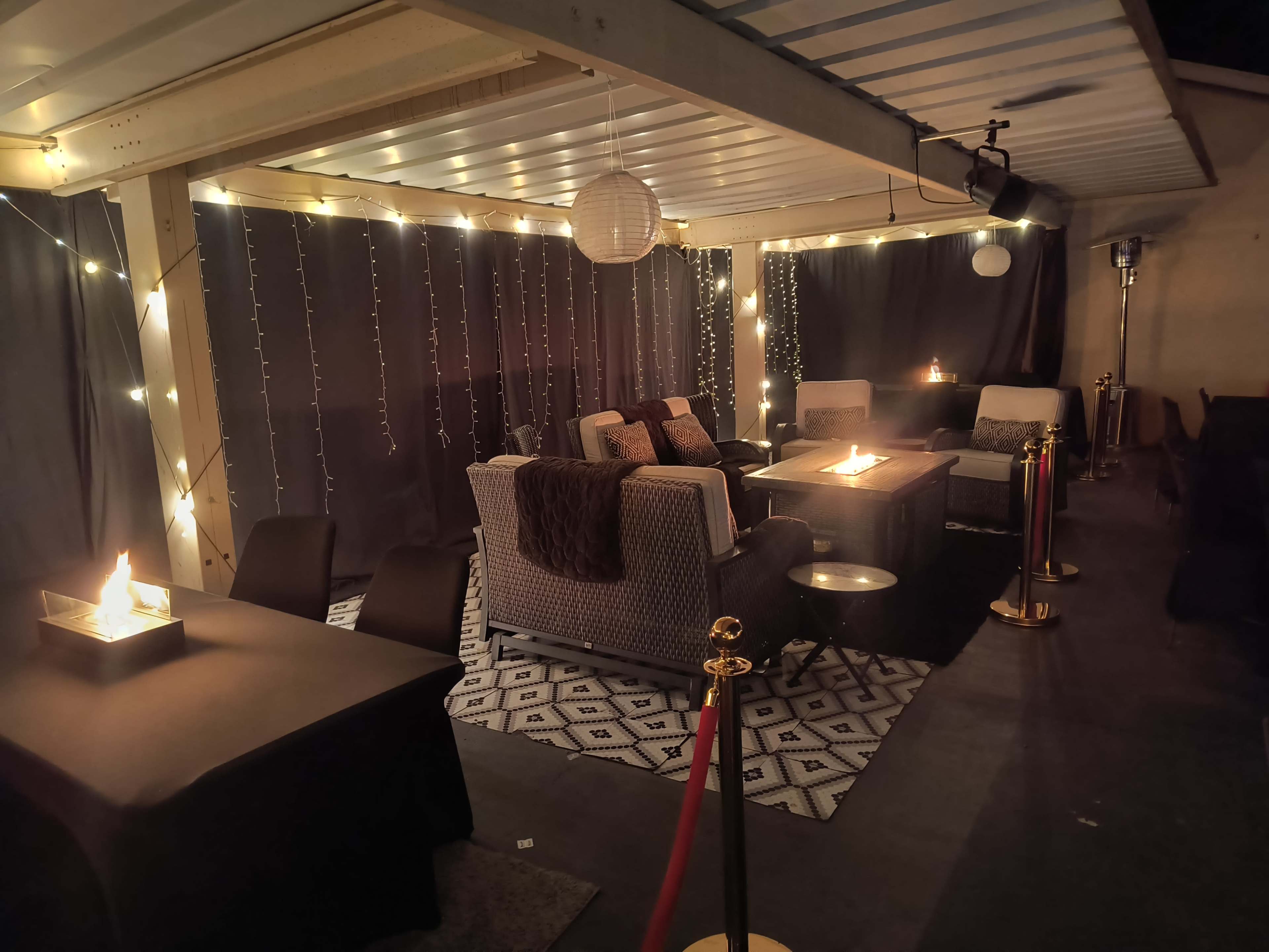 The image shows a cozy outdoor lounge area with string lights, comfortable seating, and fire pits surrounded by dark curtains.