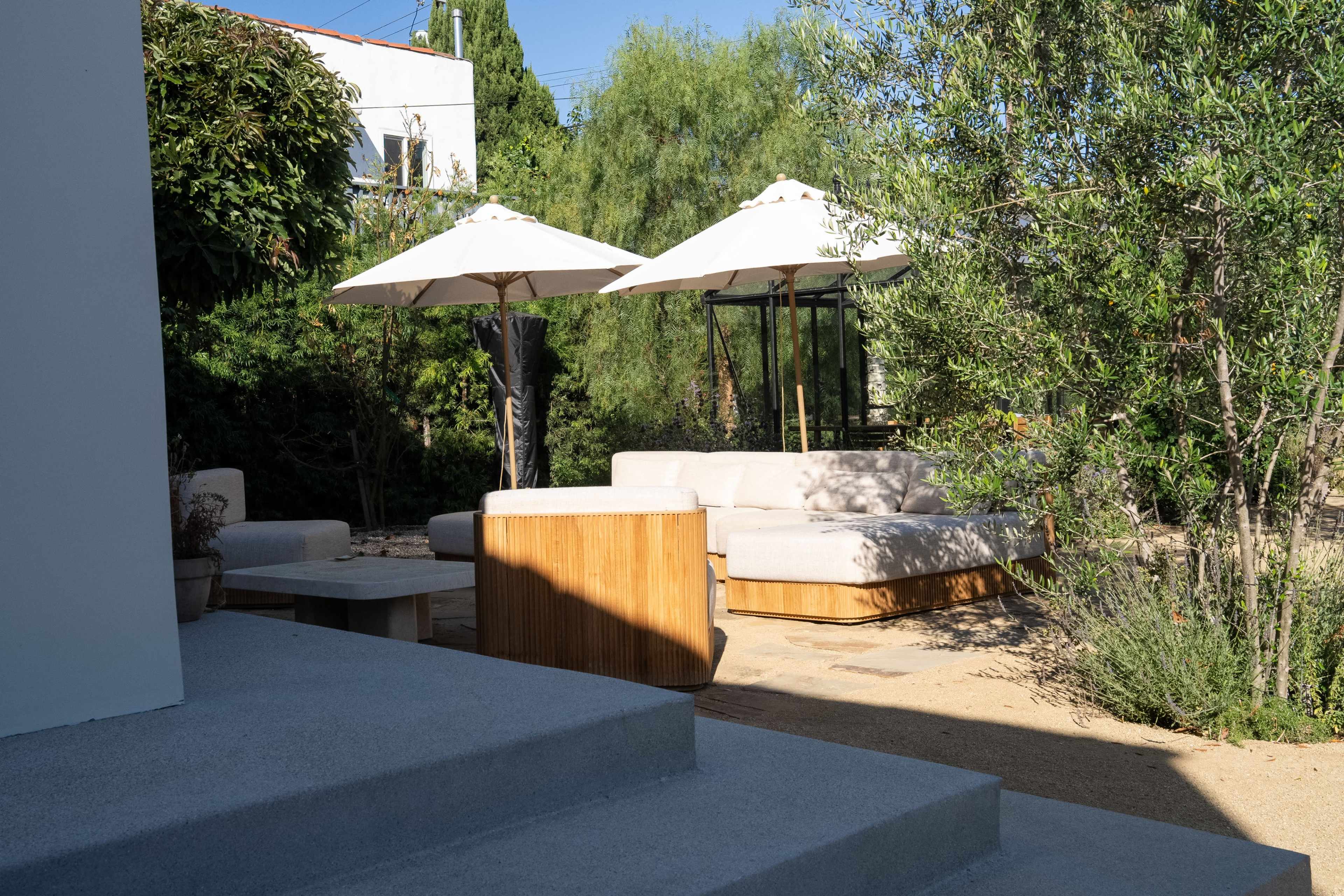 The image shows a serene outdoor seating area with a light-colored sofa, wooden table, and two umbrellas surrounded by greenery.