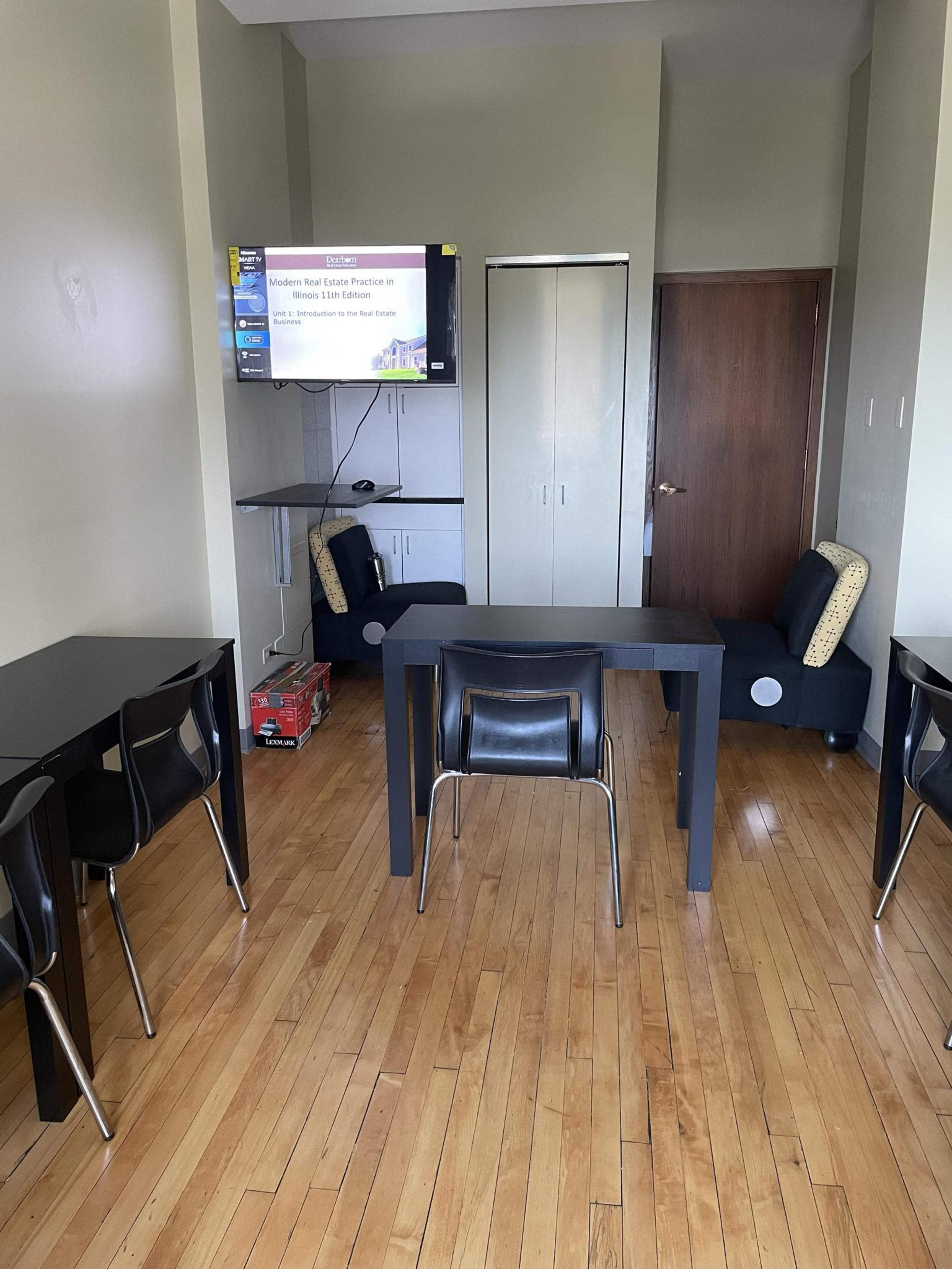 The image shows a room with wooden floors, a television mounted on the wall, a closet, and several black tables with chairs arranged around them.
