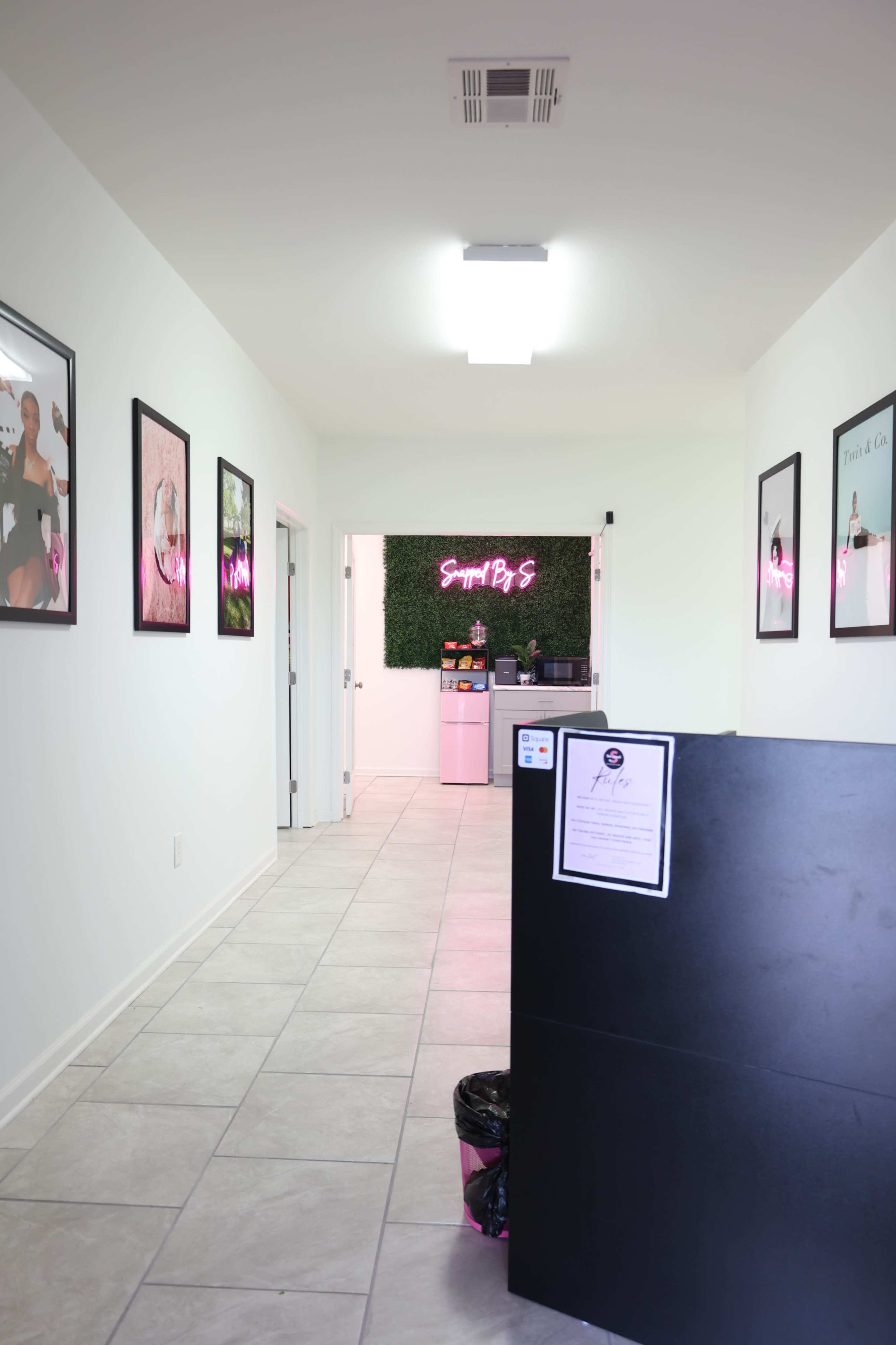 The image shows a clean hallway in an office or salon with framed pictures on the walls and a reception area at the end featuring a neon sign.