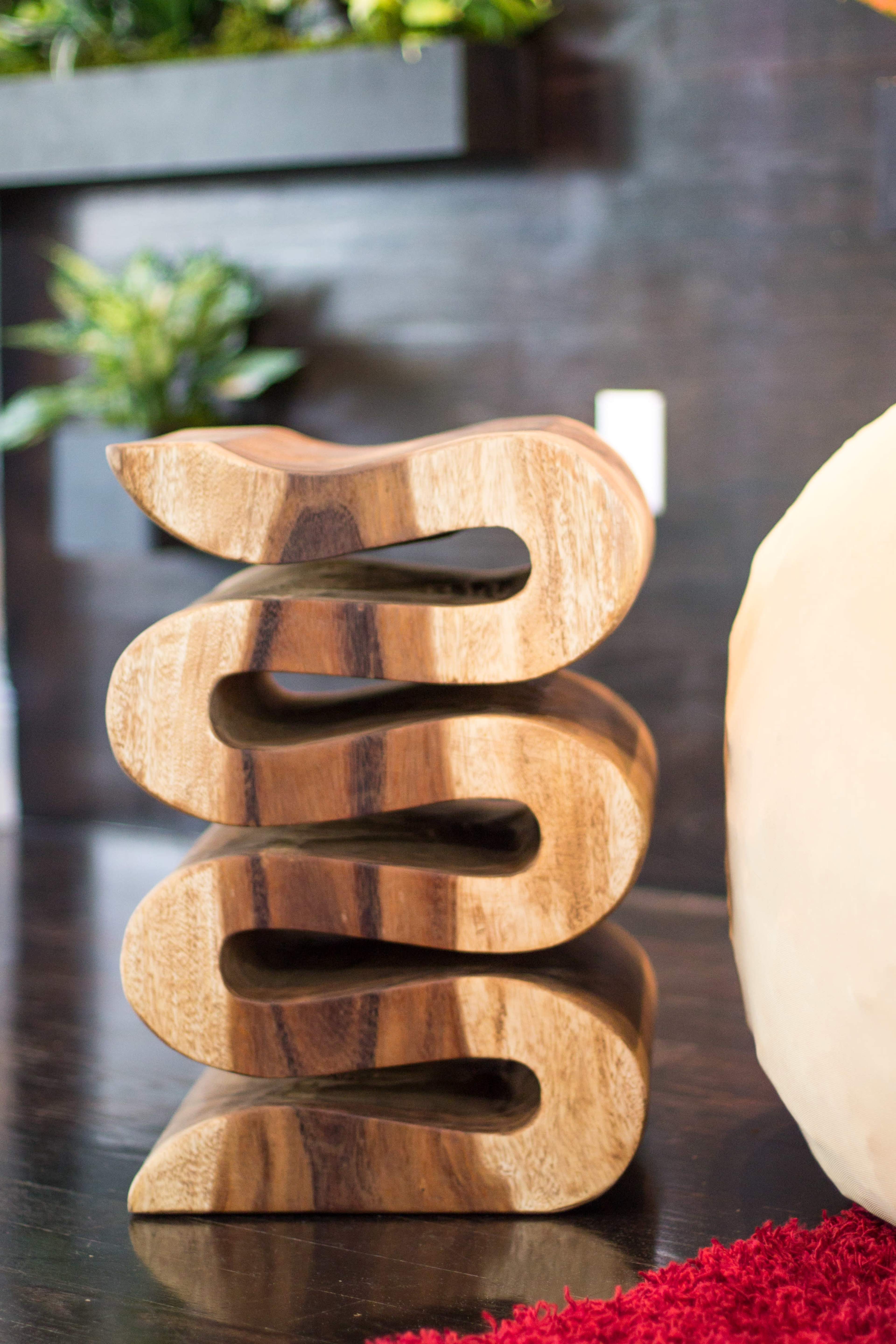 A wooden sculpture with a wavy design sits on a dark table beside a round object and a potted plant.