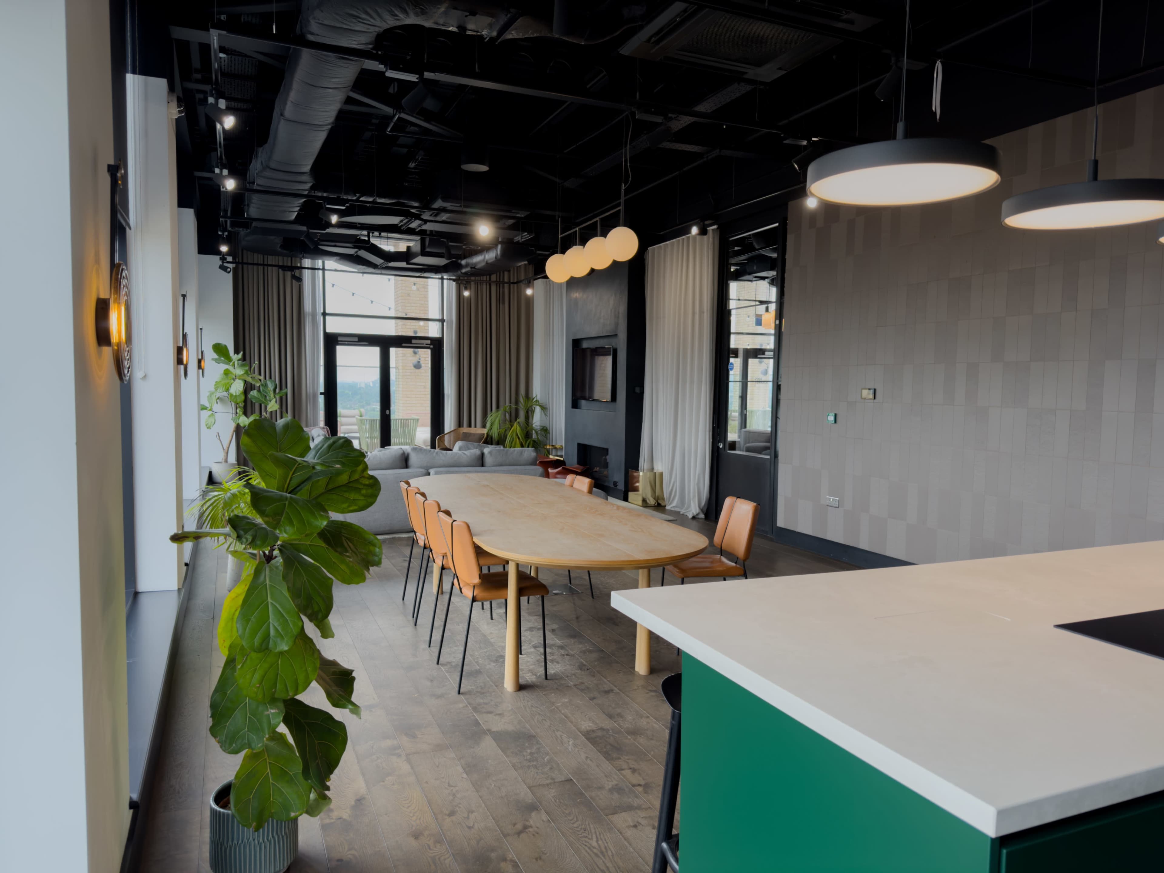 A modern office space features a large dining table with orange chairs, a lounge area with a gray sofa, and a kitchen counter with green cabinetry.