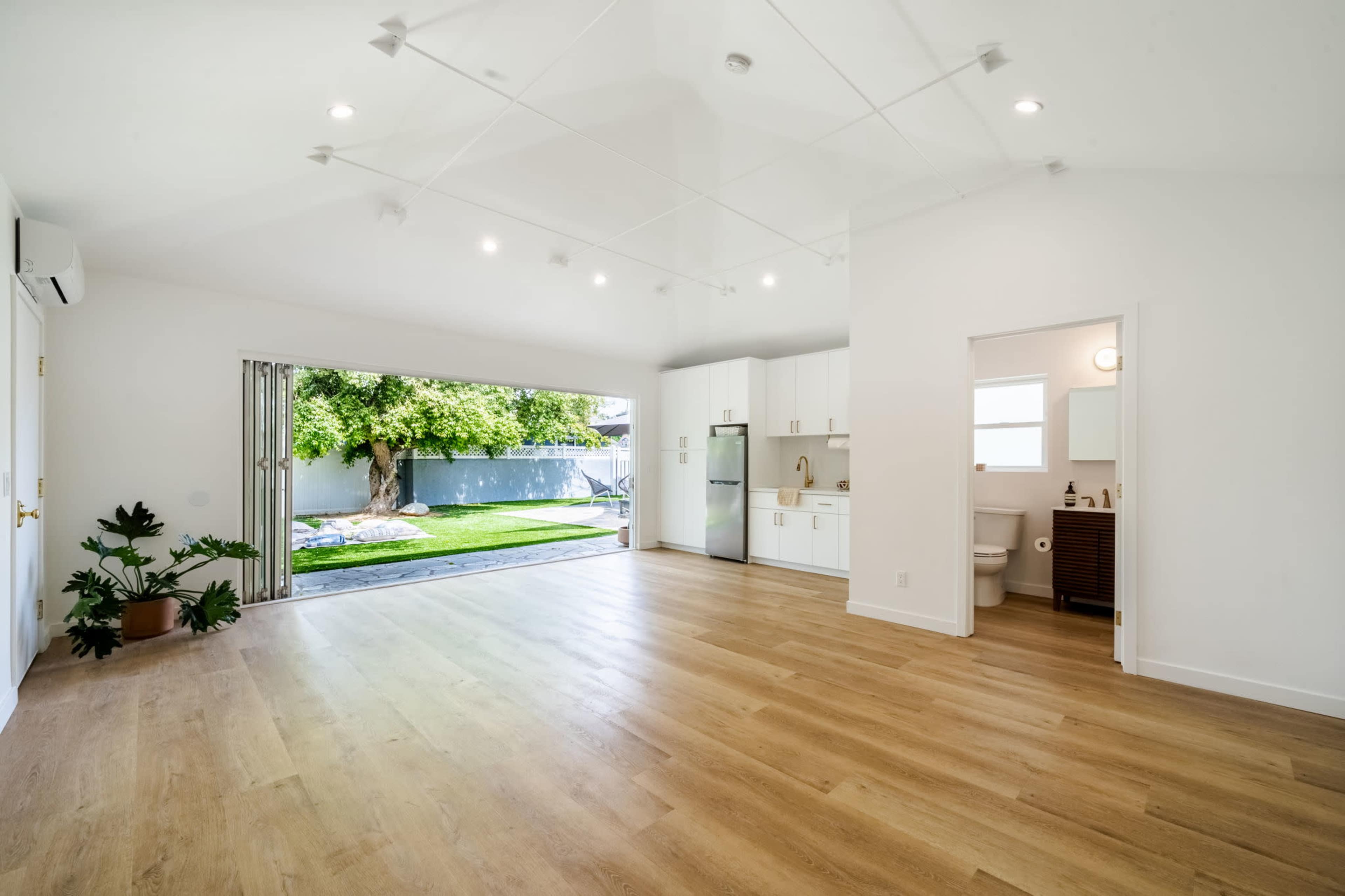 A spacious, well-lit room features an open layout with a view of a backyard, showcasing a kitchenette and a bathroom area.