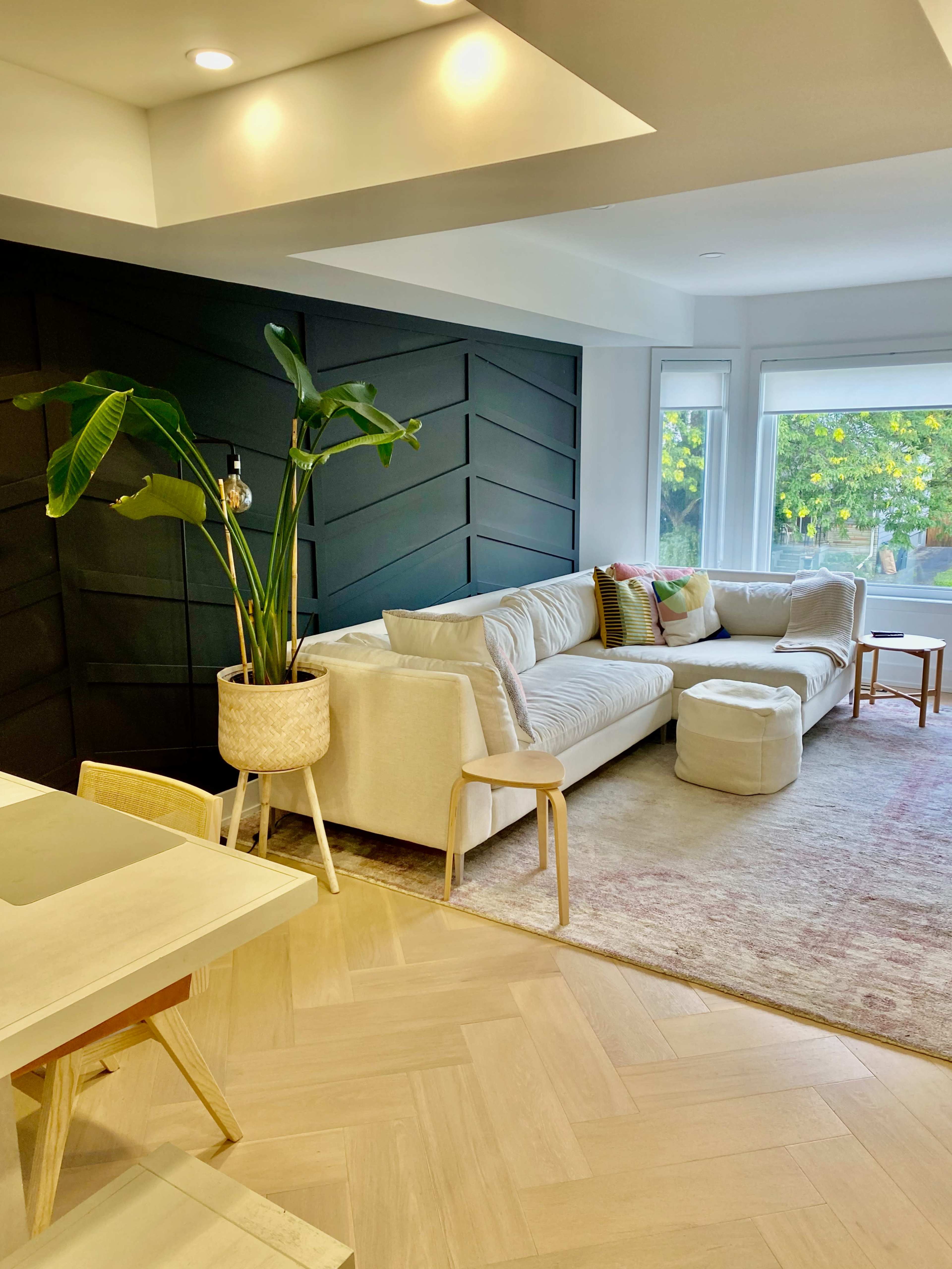 A modern living room with a light-colored sofa, a round ottoman, and a decorative plant in a corner, adjacent to a dining area with a marble table.