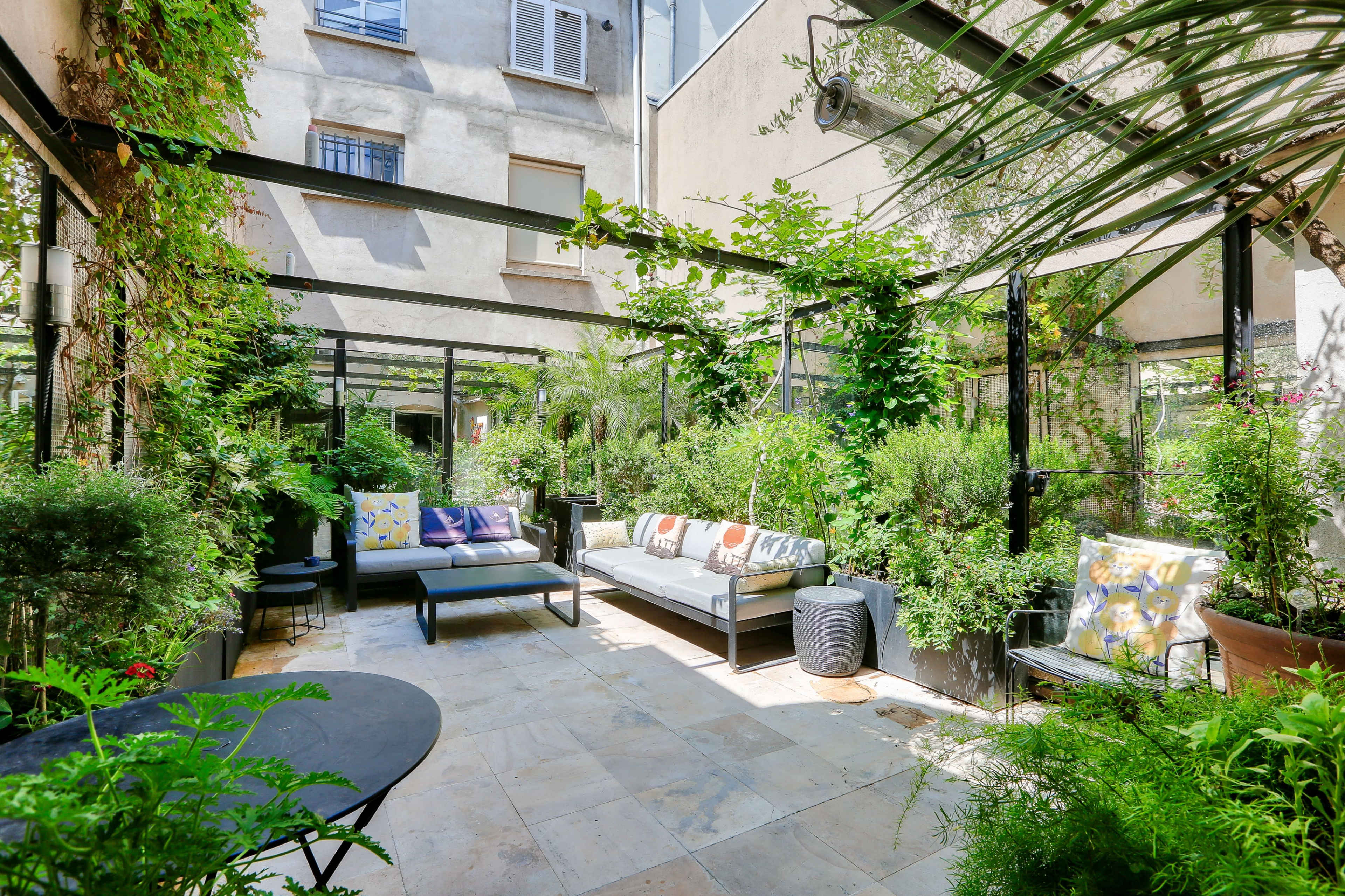 A shaded courtyard features a mix of potted plants, seating areas, and decorative pillows surrounded by greenery.