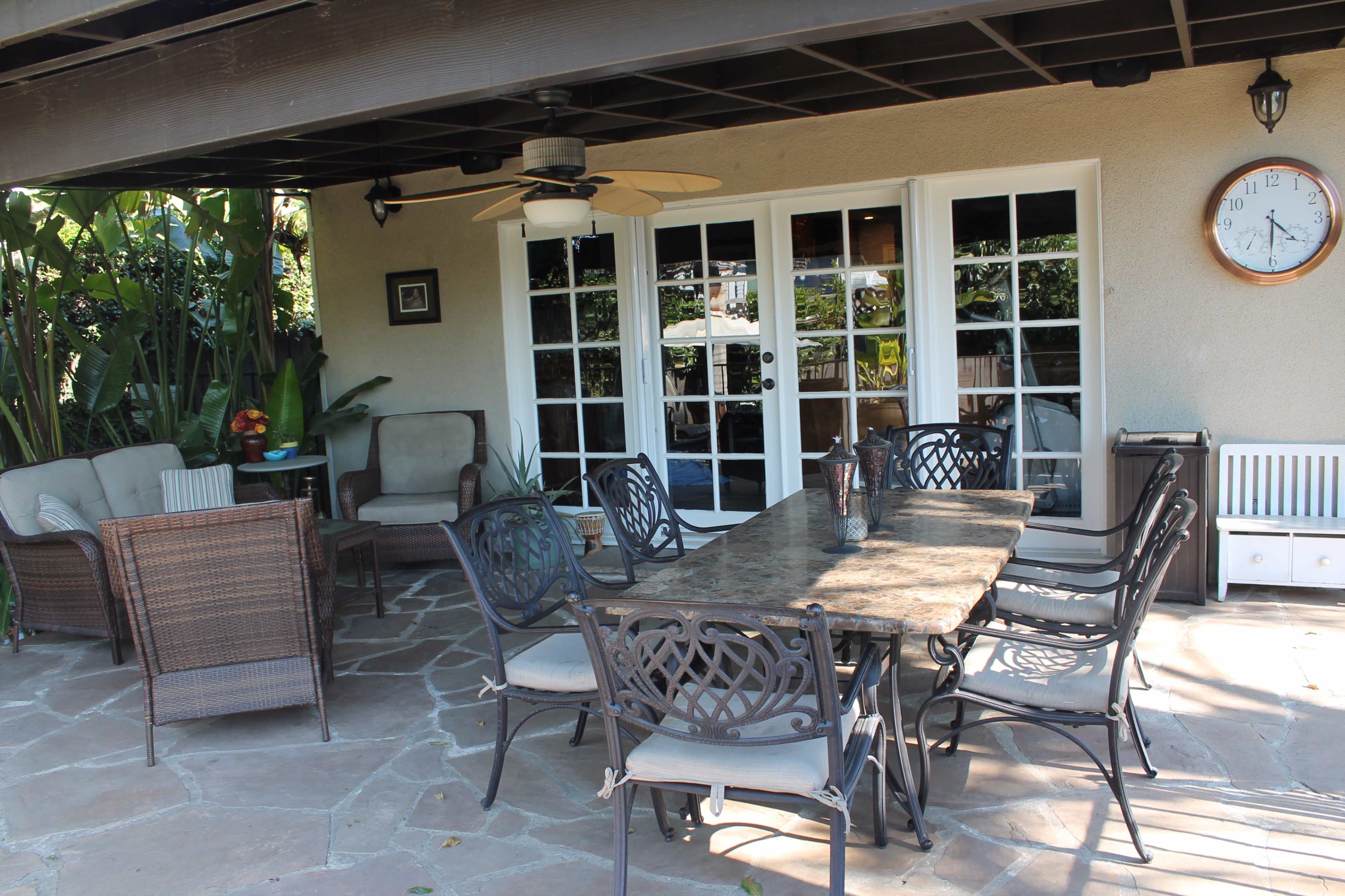 A patio area features a large stone table surrounded by metal chairs, with wicker seating and lush greenery in the background.