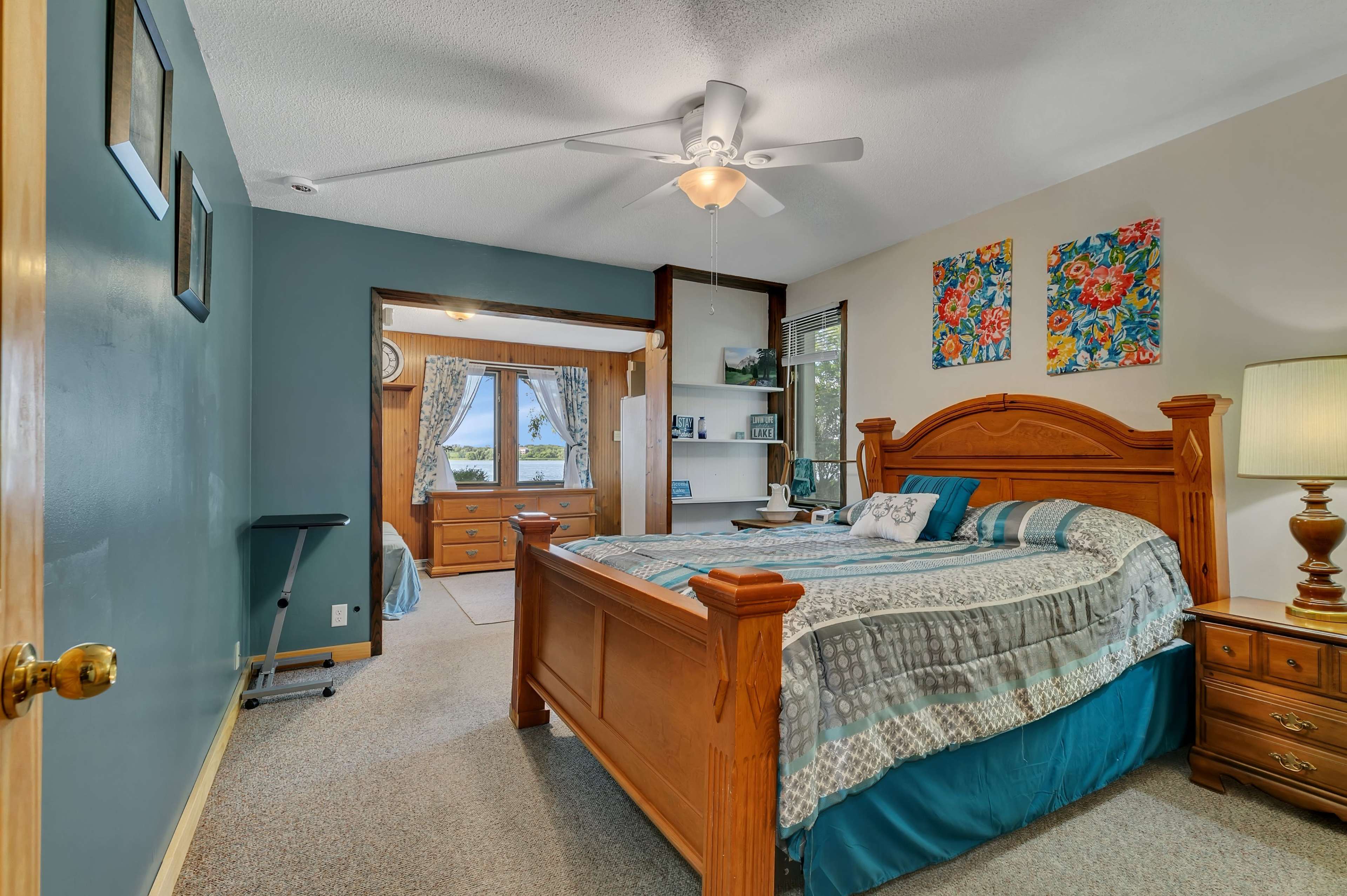 The image shows a well-furnished bedroom featuring a wooden bed with a patterned comforter, two nightstands, and a doorway leading to another room.