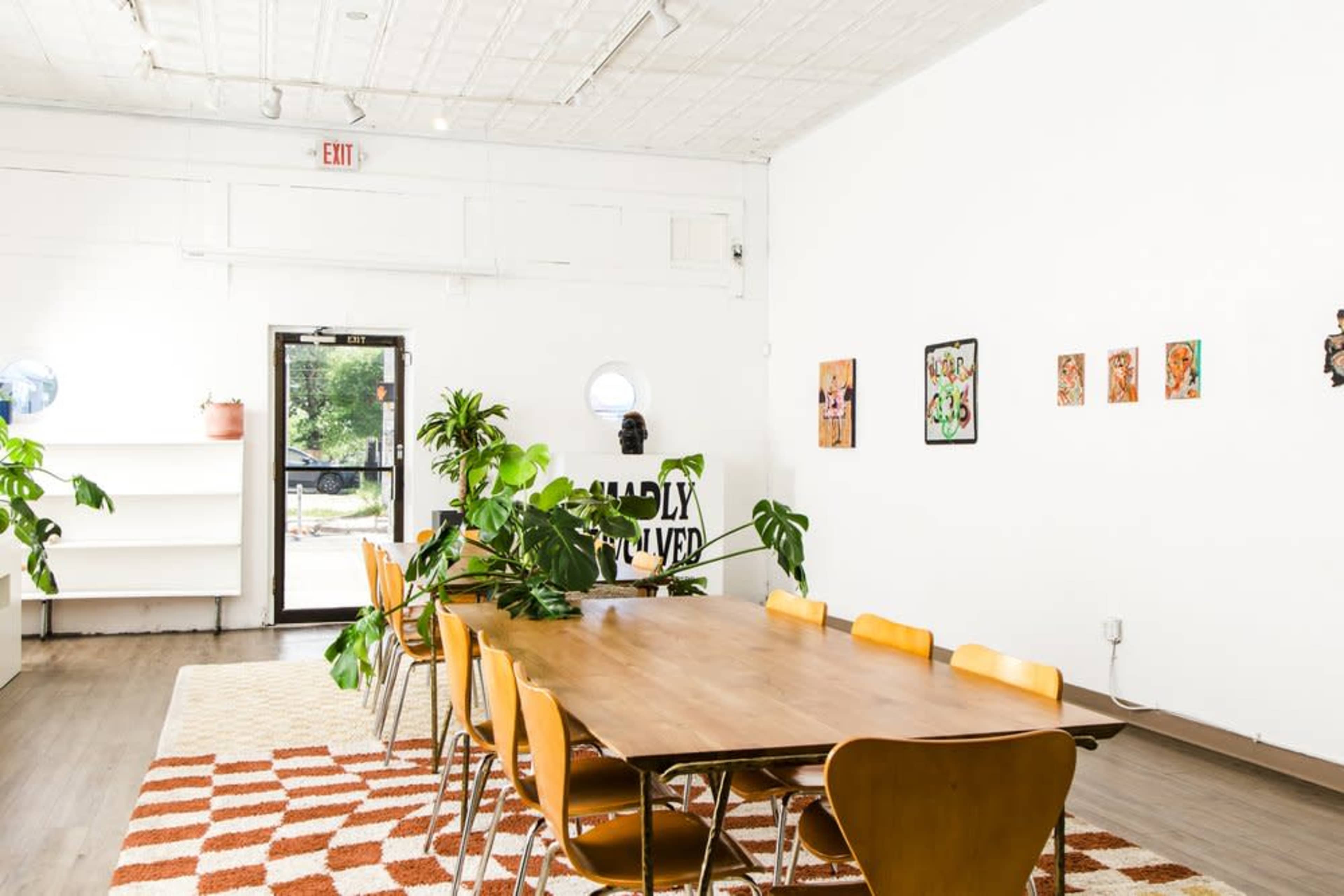 A spacious room features a wooden dining table surrounded by yellow chairs, adorned with plants and wall art, with a doorway leading outside.