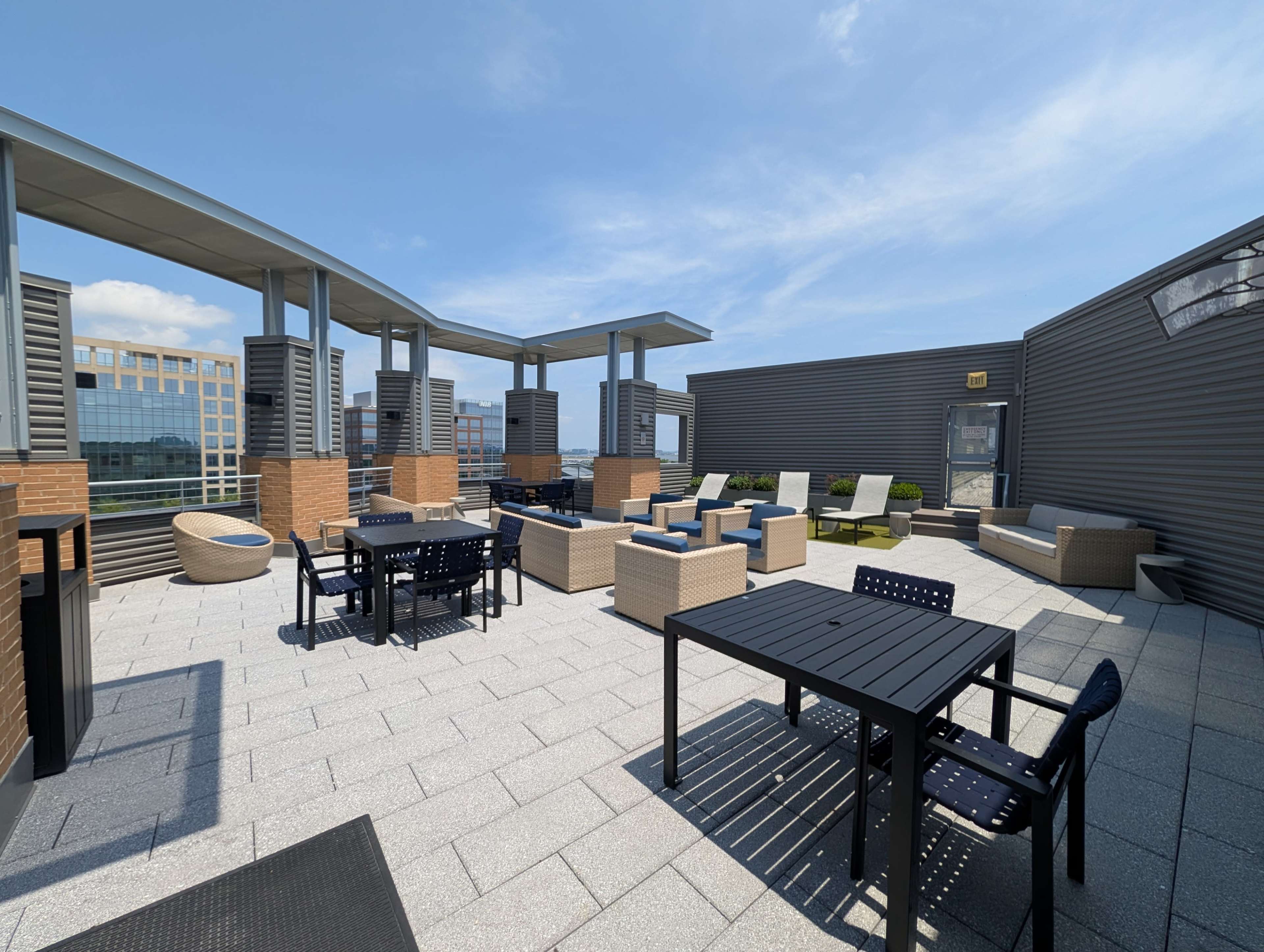 The image shows a spacious rooftop terrace with seating areas featuring tables, chairs, and lounge furniture, surrounded by a modern architectural backdrop.