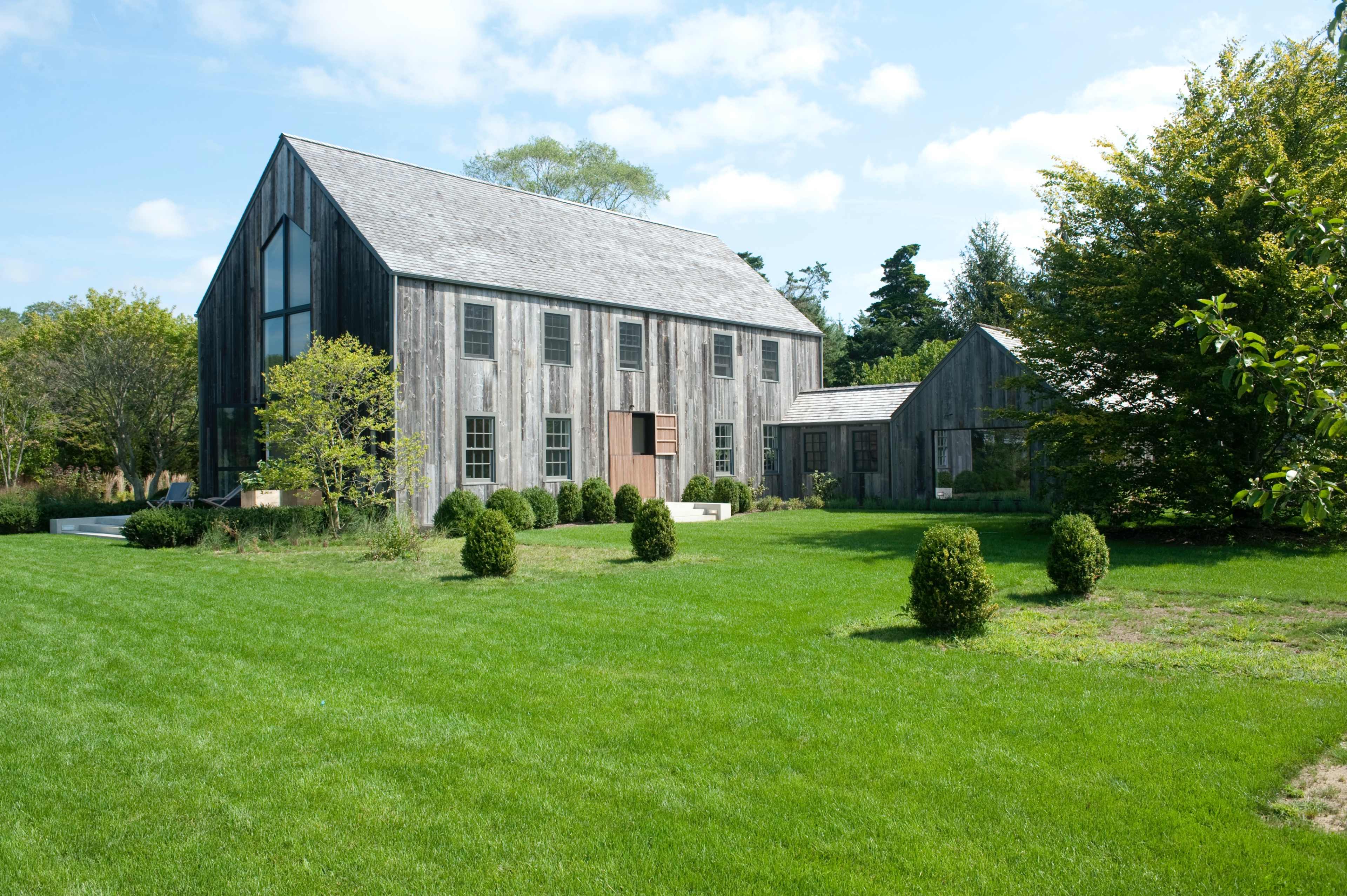 A large, contemporary barn-style building made of weathered wood is set on a green lawn surrounded by neatly trimmed bushes and trees.