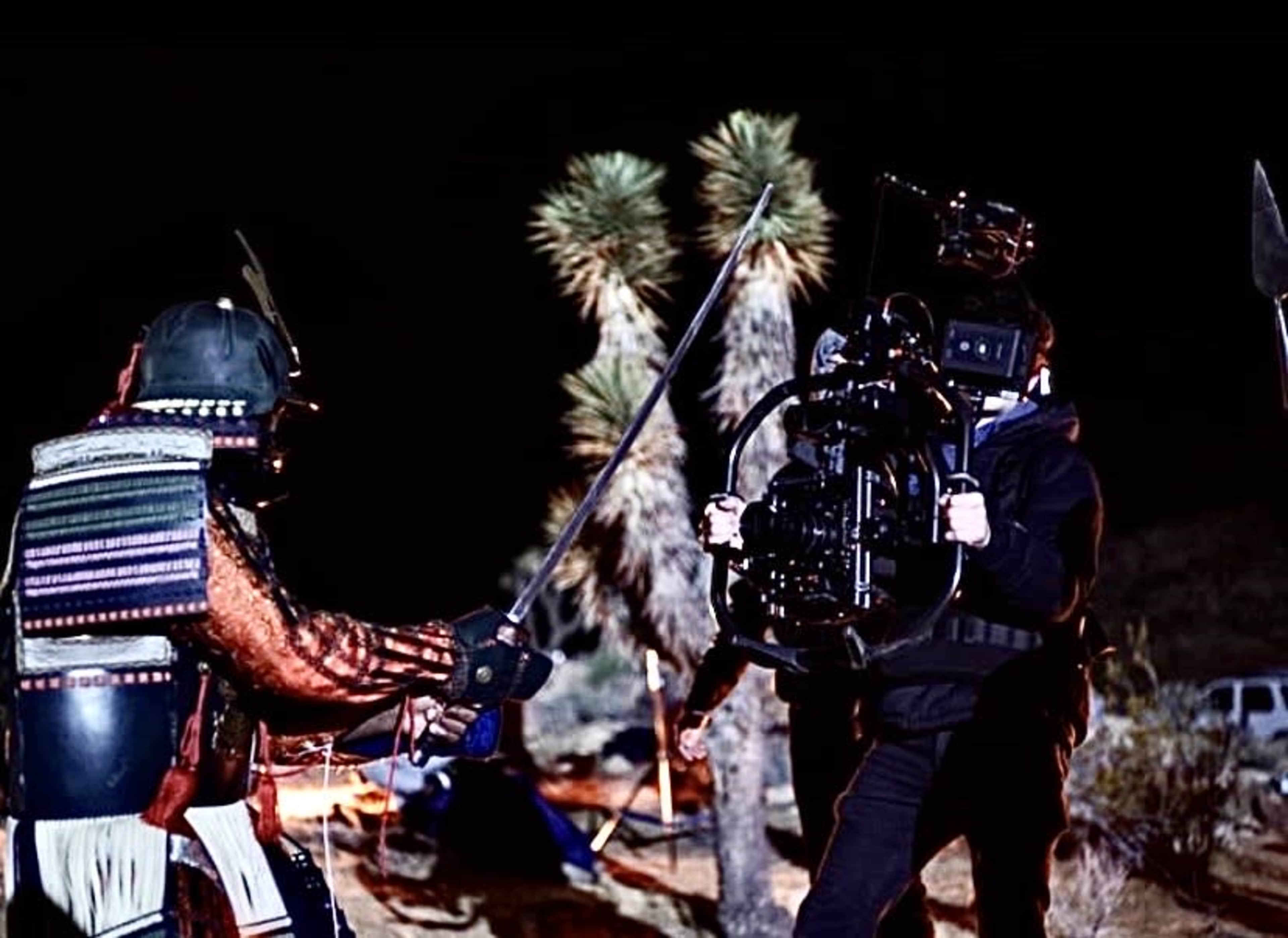 A crew member operates a camera while two individuals dressed in traditional attire engage in a mock battle scene in a desert setting at night.
