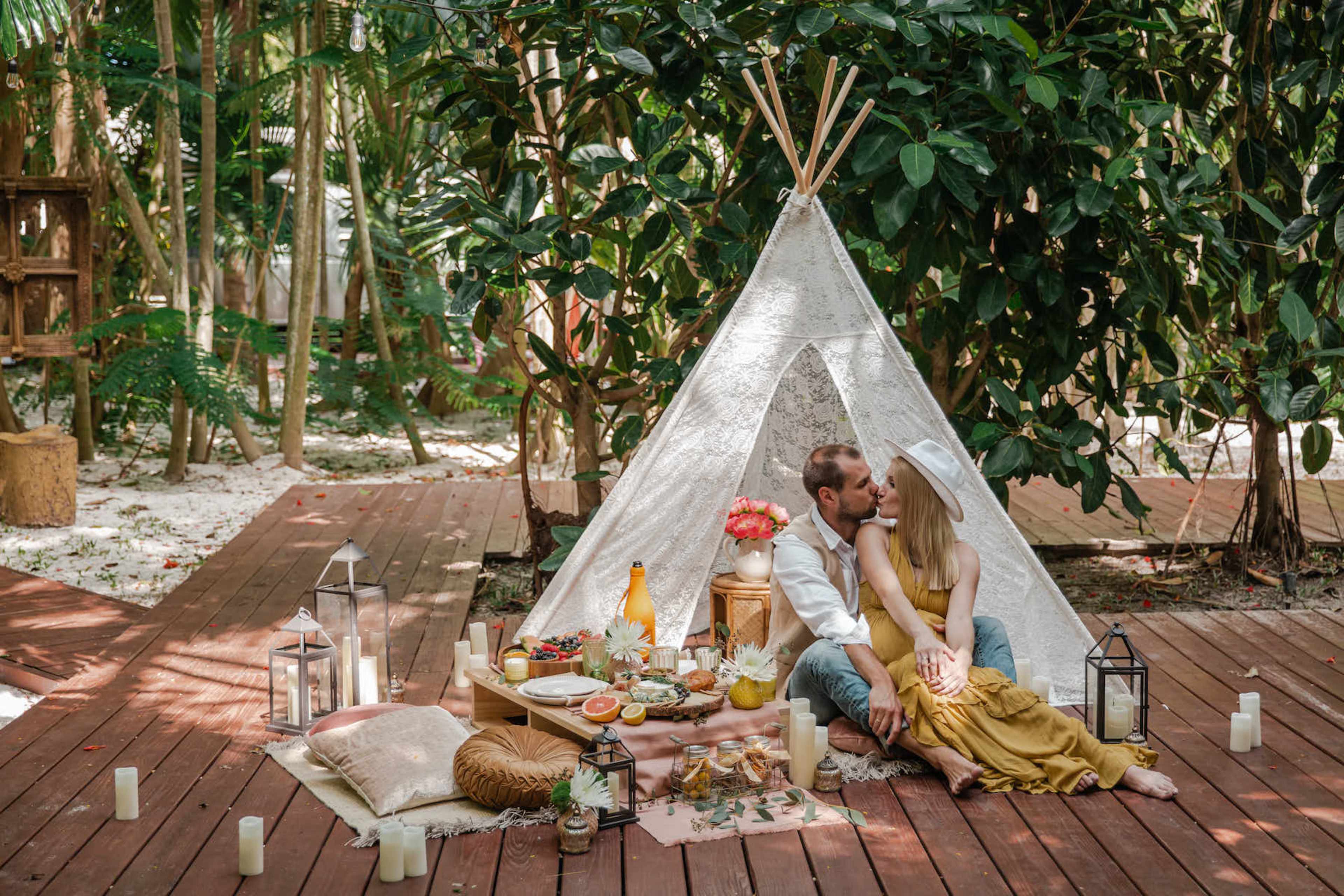 A couple shares an intimate moment in front of a decorated teepee on a wooden deck surrounded by greenery, with a picnic spread laid out nearby.