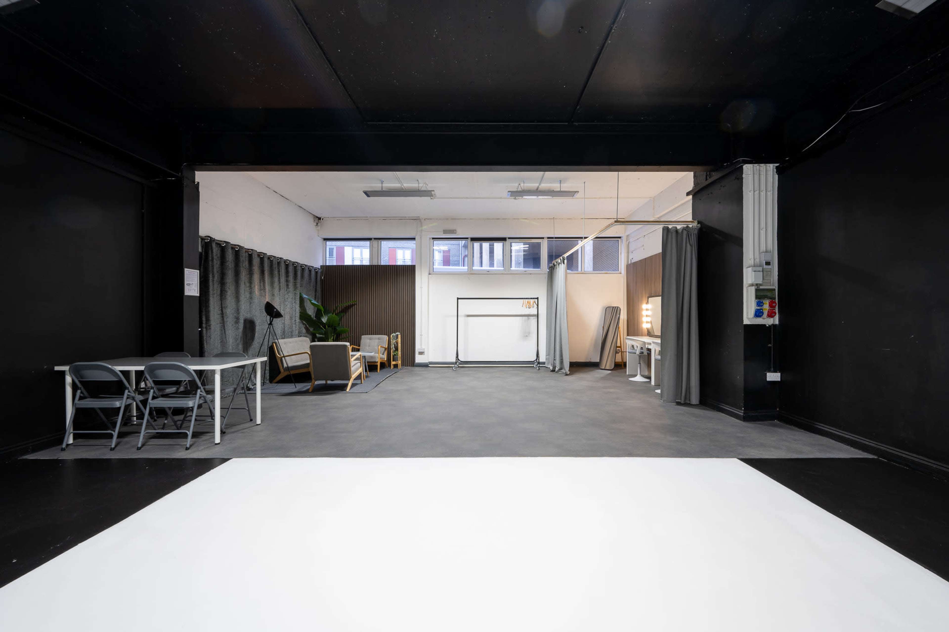 The image depicts a spacious, empty studio with black walls and a white floor, featuring a few pieces of furniture, including a table and chairs, and a potted plant.